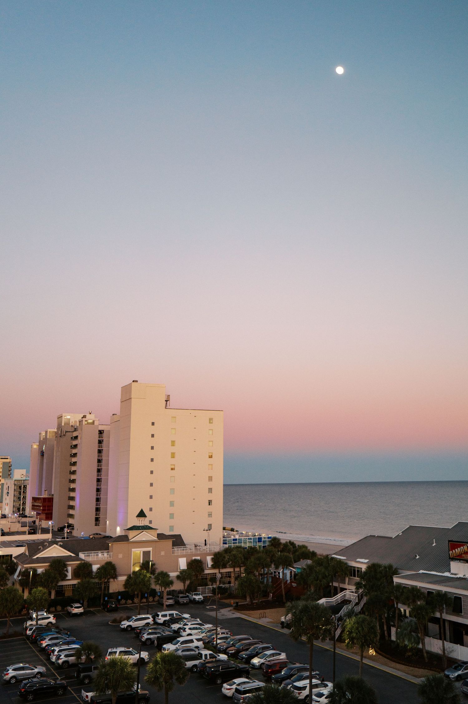 Beachfront buildings and hotels line the coast at dusk with a full moon rising in the pale blue sky.