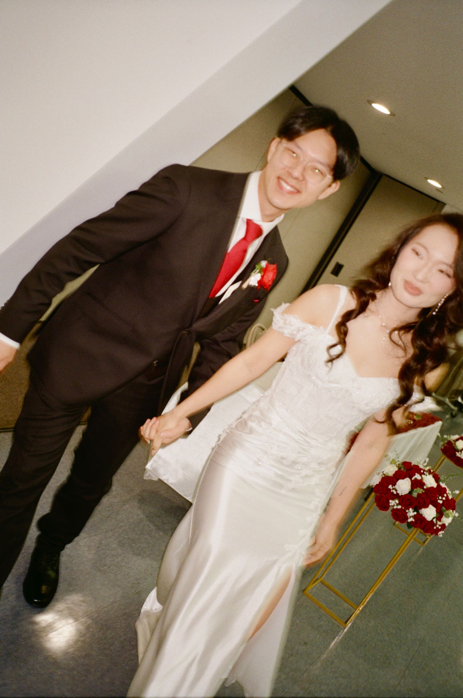 A wedding couple poses in a reception hall with red floral bouquet accents.