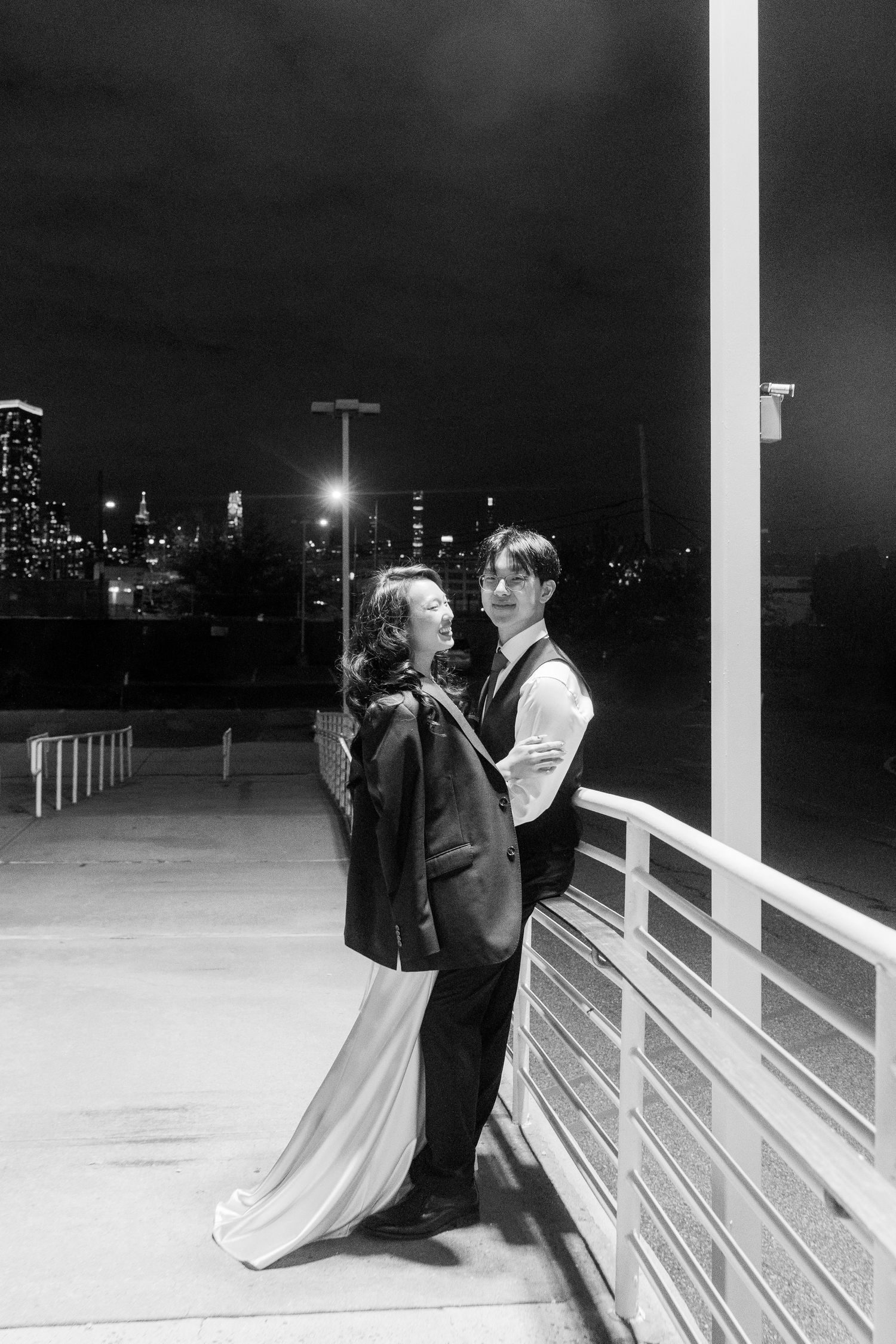 Romantic nighttime couple portrait series with city lights in background against white railings.