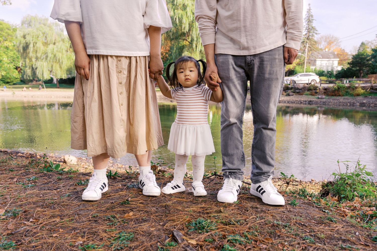 Family wearing coordinated neutral outfits standing by a pond in autumn.