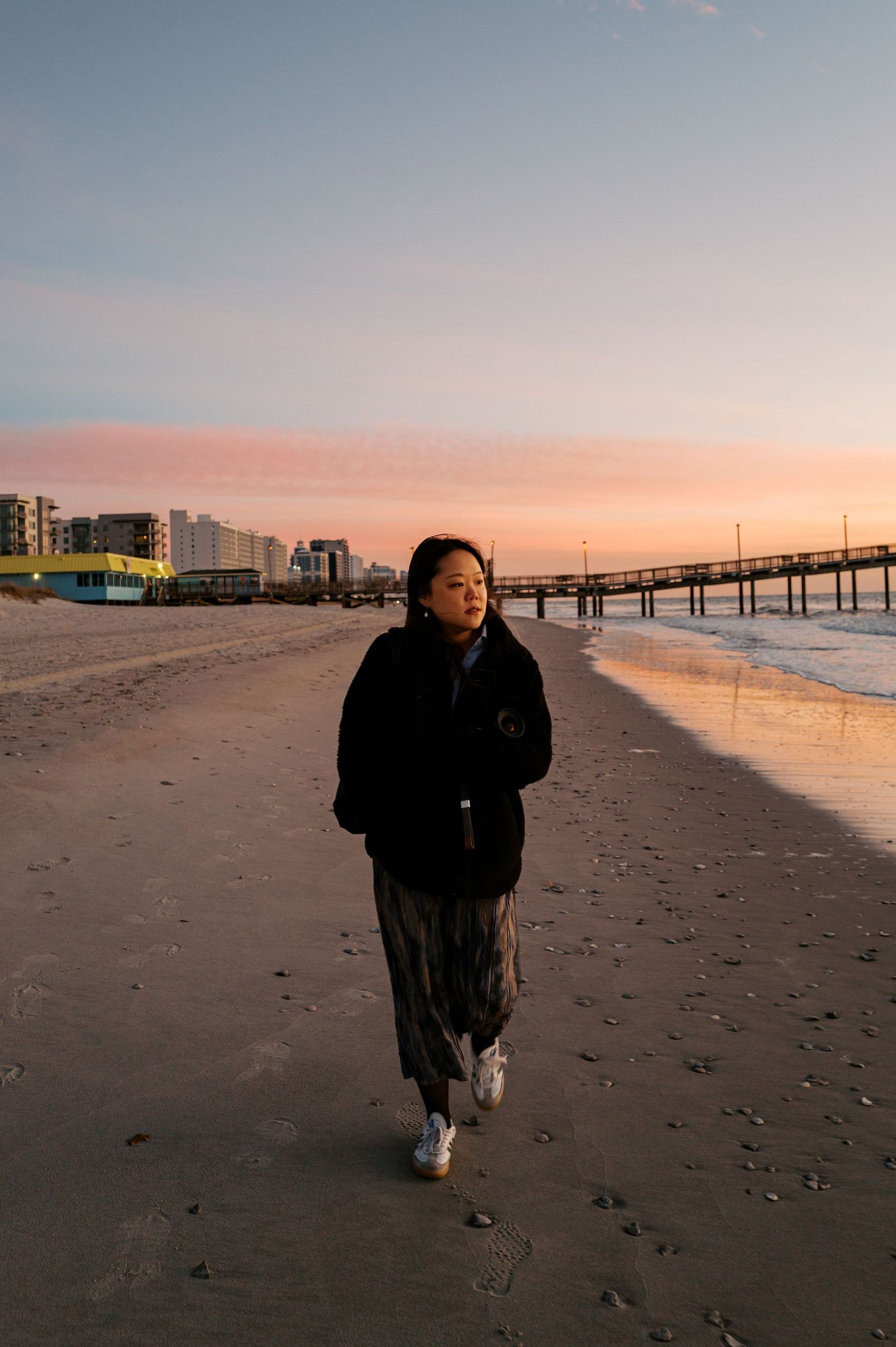 A solitary figure walks along a sandy beach at sunset with a pier visible in the distance.
