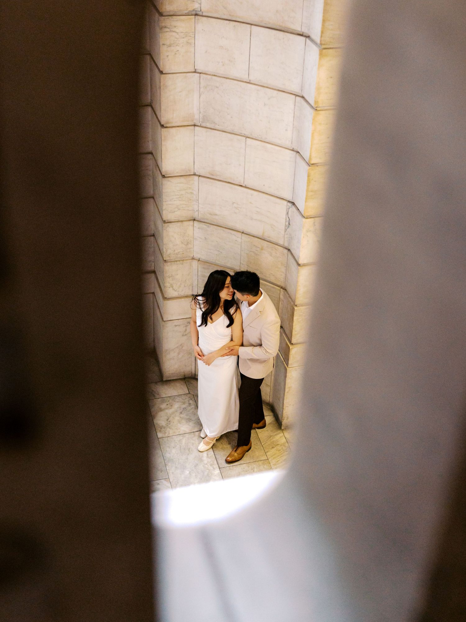 Couple sharing a romantic moment in a stone archway, dressed in white and beige outfits for an engagement photo.