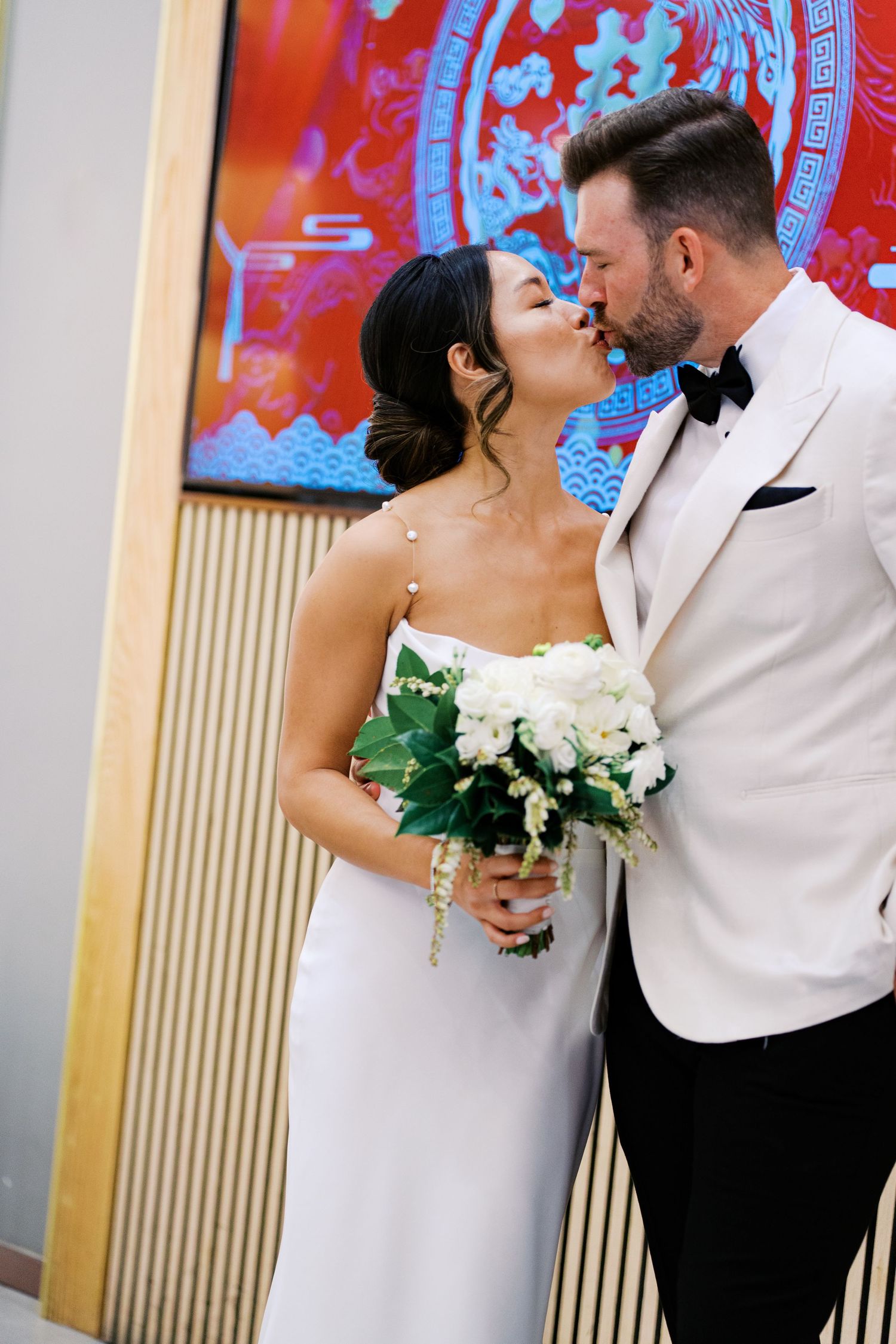 Newlywed couple shares romantic kisses against a vibrant red and yellow backdrop during their wedding photo session.