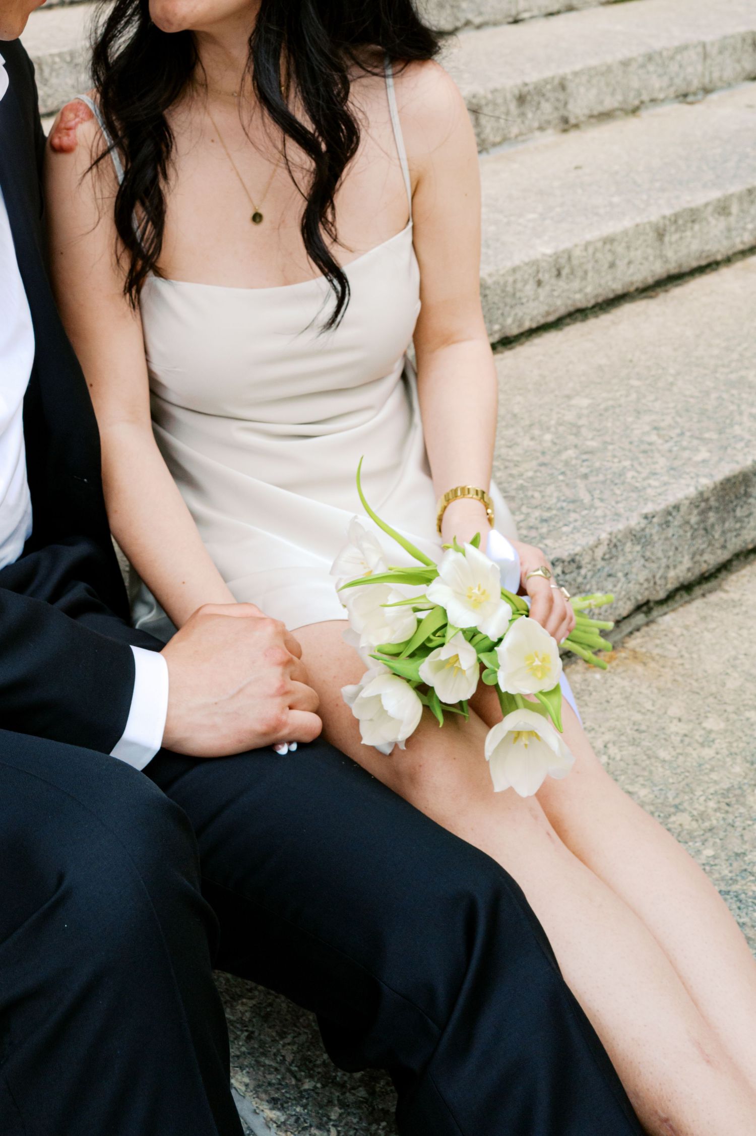 Bride in ivory slip dress holding white tulip bouquet sits on stone steps beside groom in navy suit.