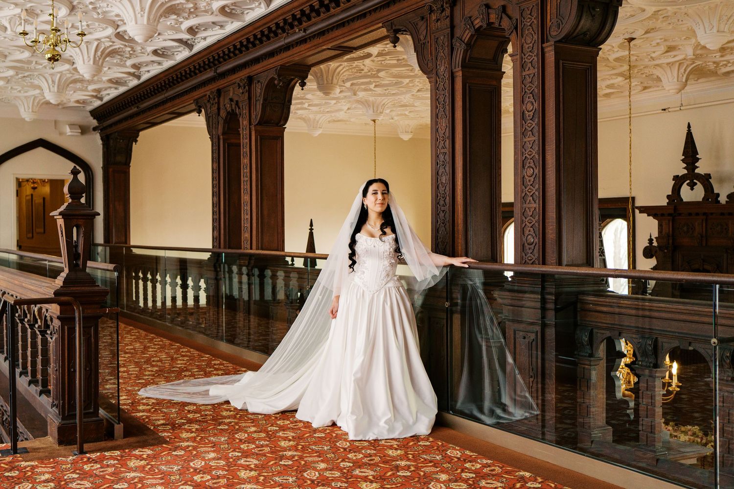 A bride in a white wedding gown stands in an elegant historic mansion with ornate wooden banisters and marble floors.