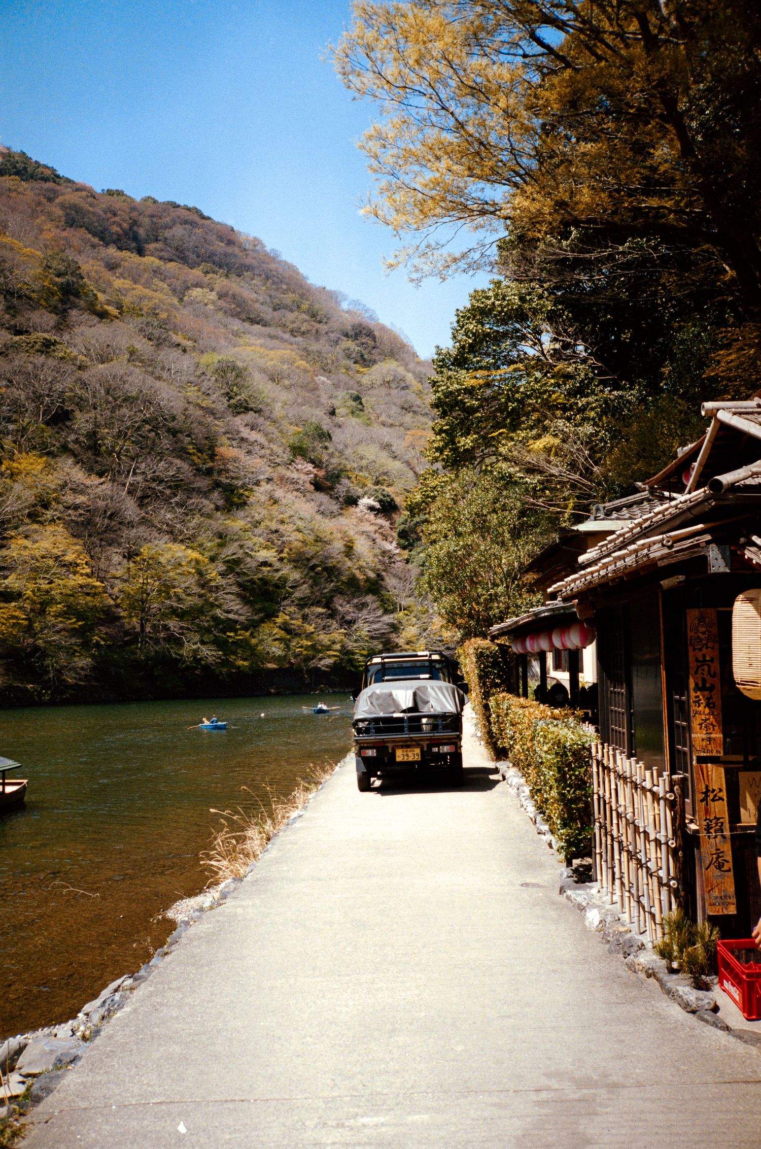 Traditional Japanese riverside path with parked car and autumn trees along water's edge.