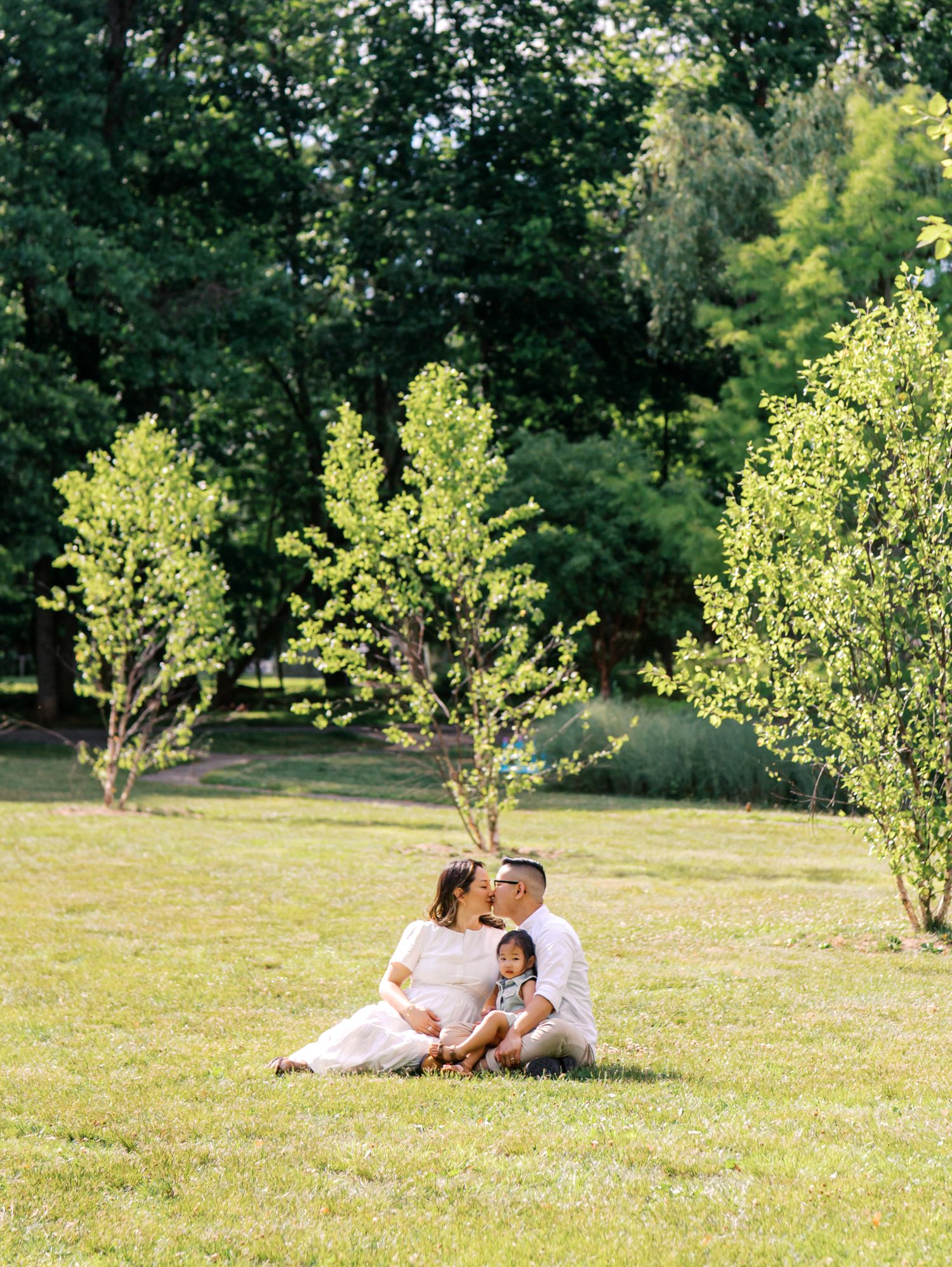 A couple sits on green grass kissing, with a toddler between them, surrounded by lush trees in a sunny park.
