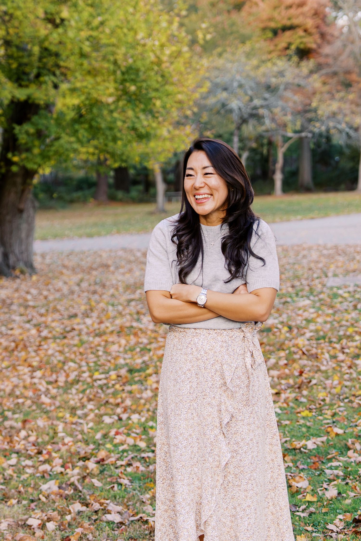 Woman in polka dot dress poses joyfully against autumn trees and fallen leaves.