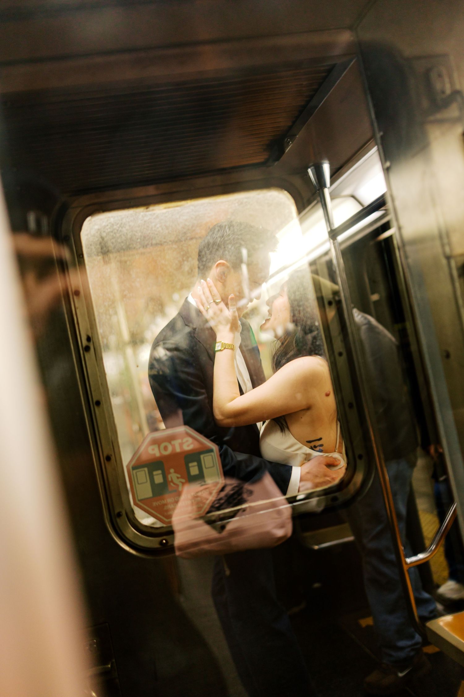 Couple sharing a romantic kiss on a subway train, captured through the window in warm light.