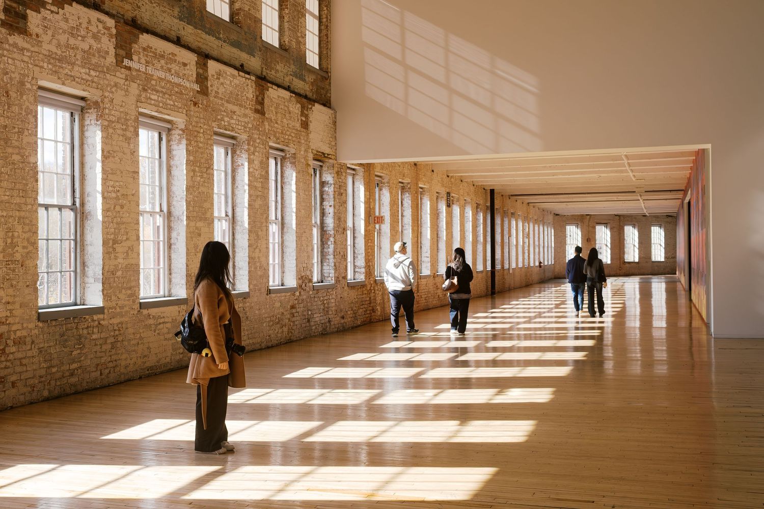 Sunlight streams through tall windows in a historic brick building hallway where people stand in the warm glow.