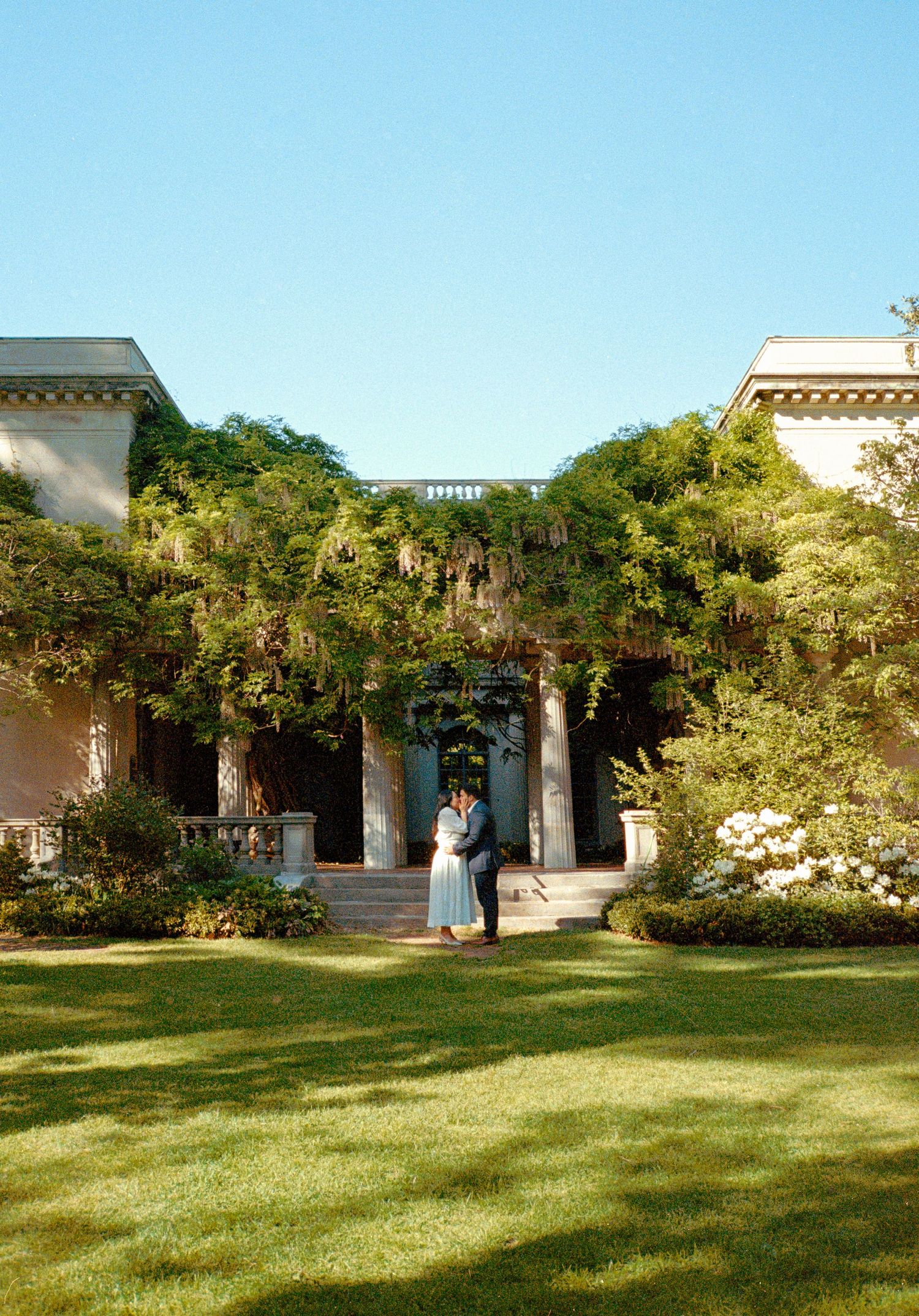 A couple stands in a lush garden setting with white columns and green lawn during a sunny outdoor photoshoot.