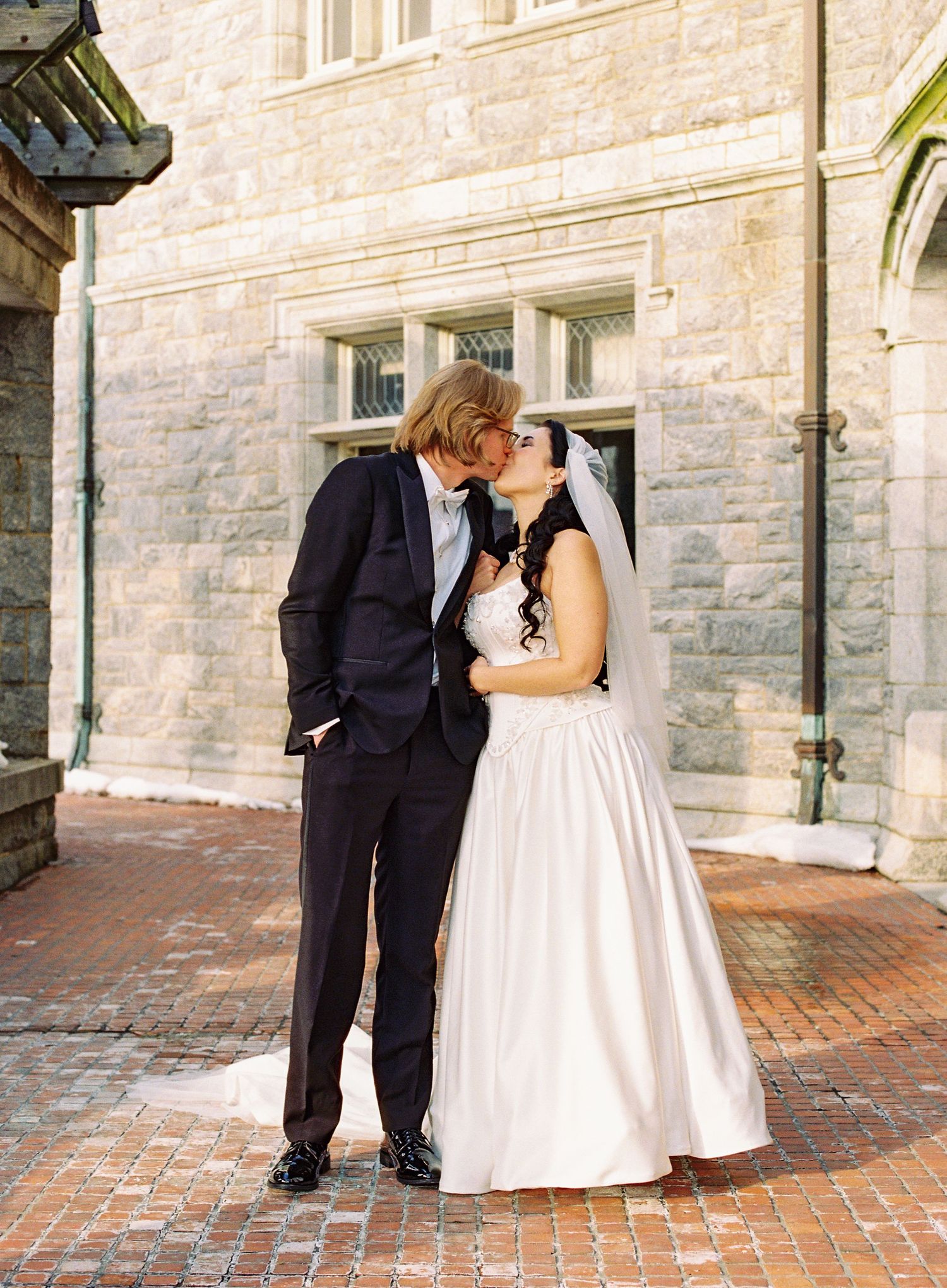 A couple shares a romantic kiss on a brick patio outside a stone building during their wedding celebration.
