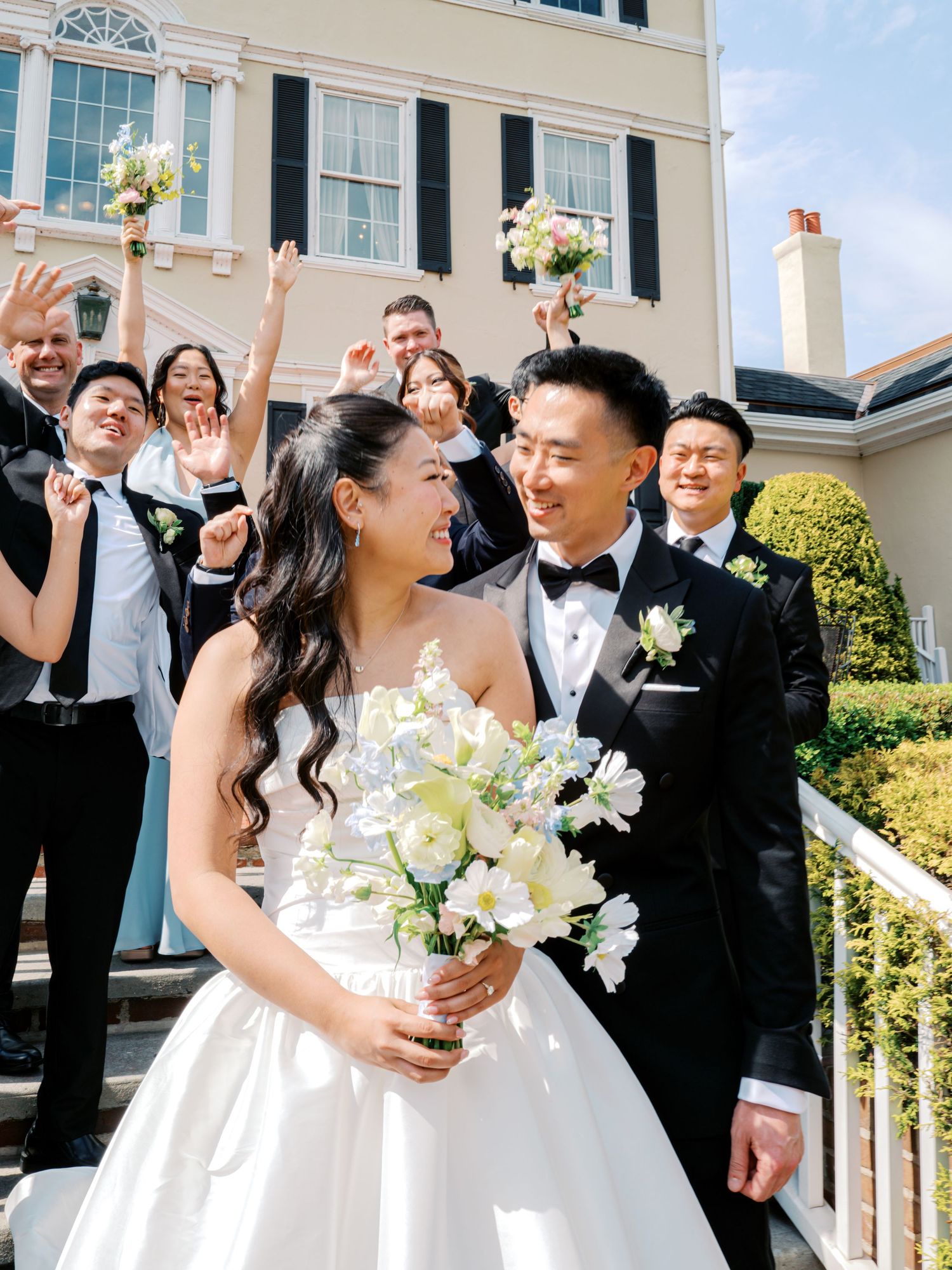 Asian bride and groom share a joyful moment on steps while wedding party celebrates behind them at elegant mansion.