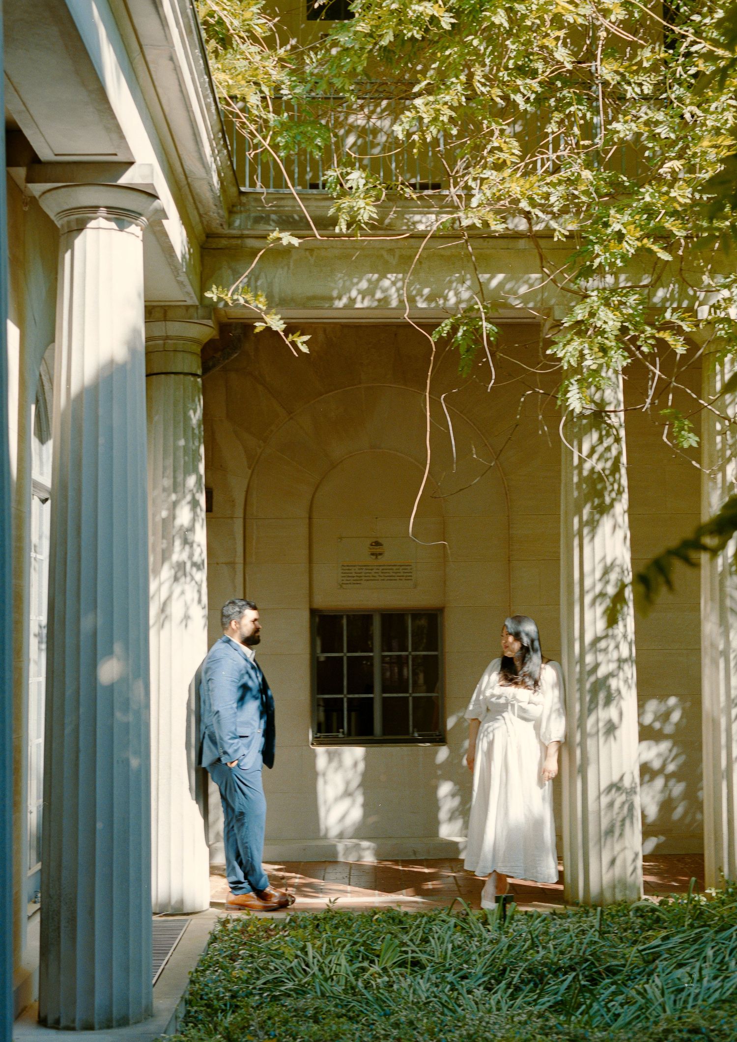 A covered walkway with white columns and greenery along a historic building.
