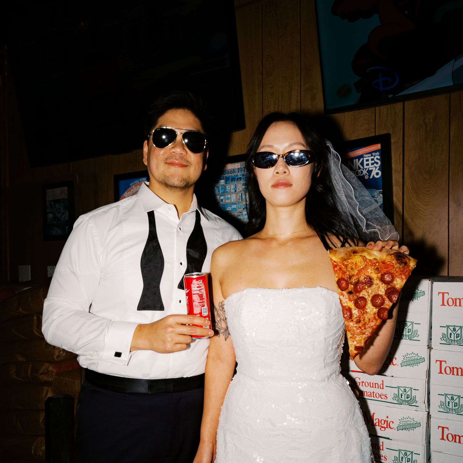A couple in wedding attire poses casually near stacked pizza boxes in a diner.