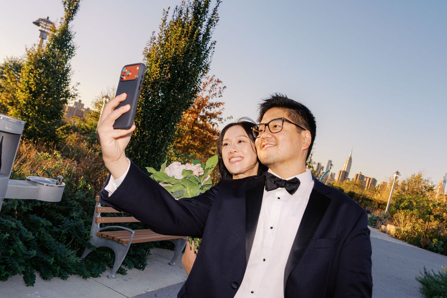 Couple in formal attire taking selfies against autumn trees.