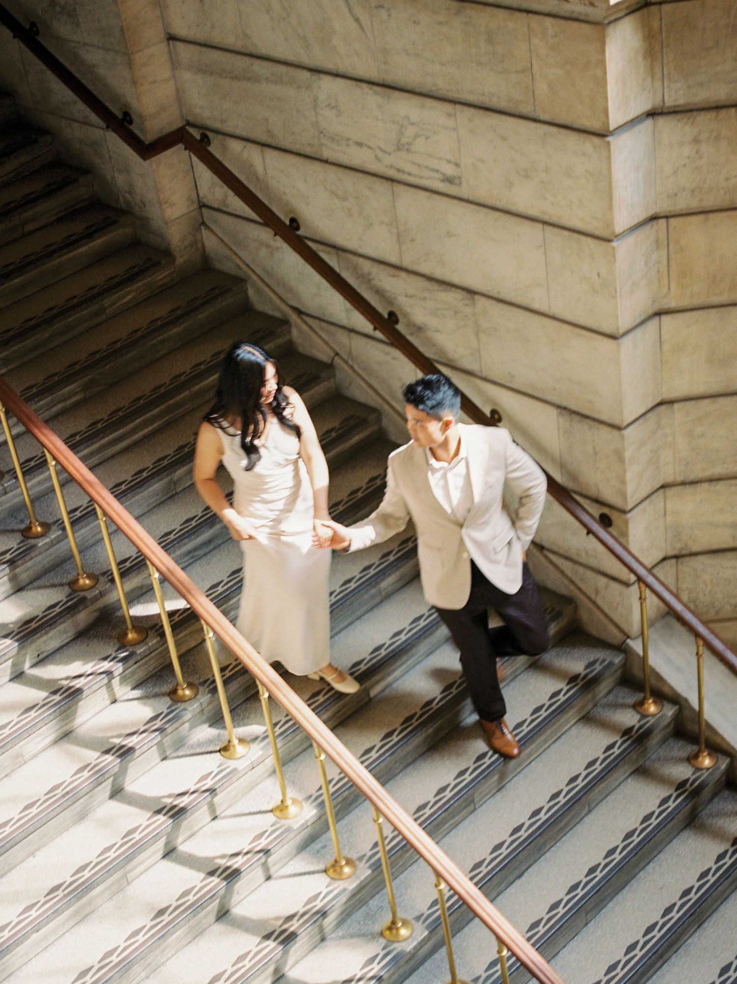 Couple holding hands walking up elegant marble staircase with brass railings in a grand building.