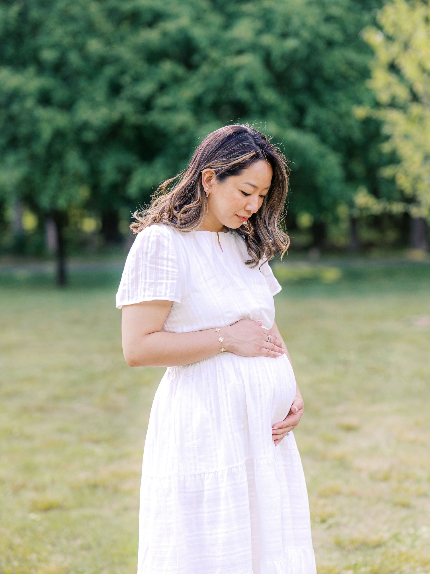 Pregnant woman in white dress cradling baby bump outdoors in a sunlit park with lush green trees.