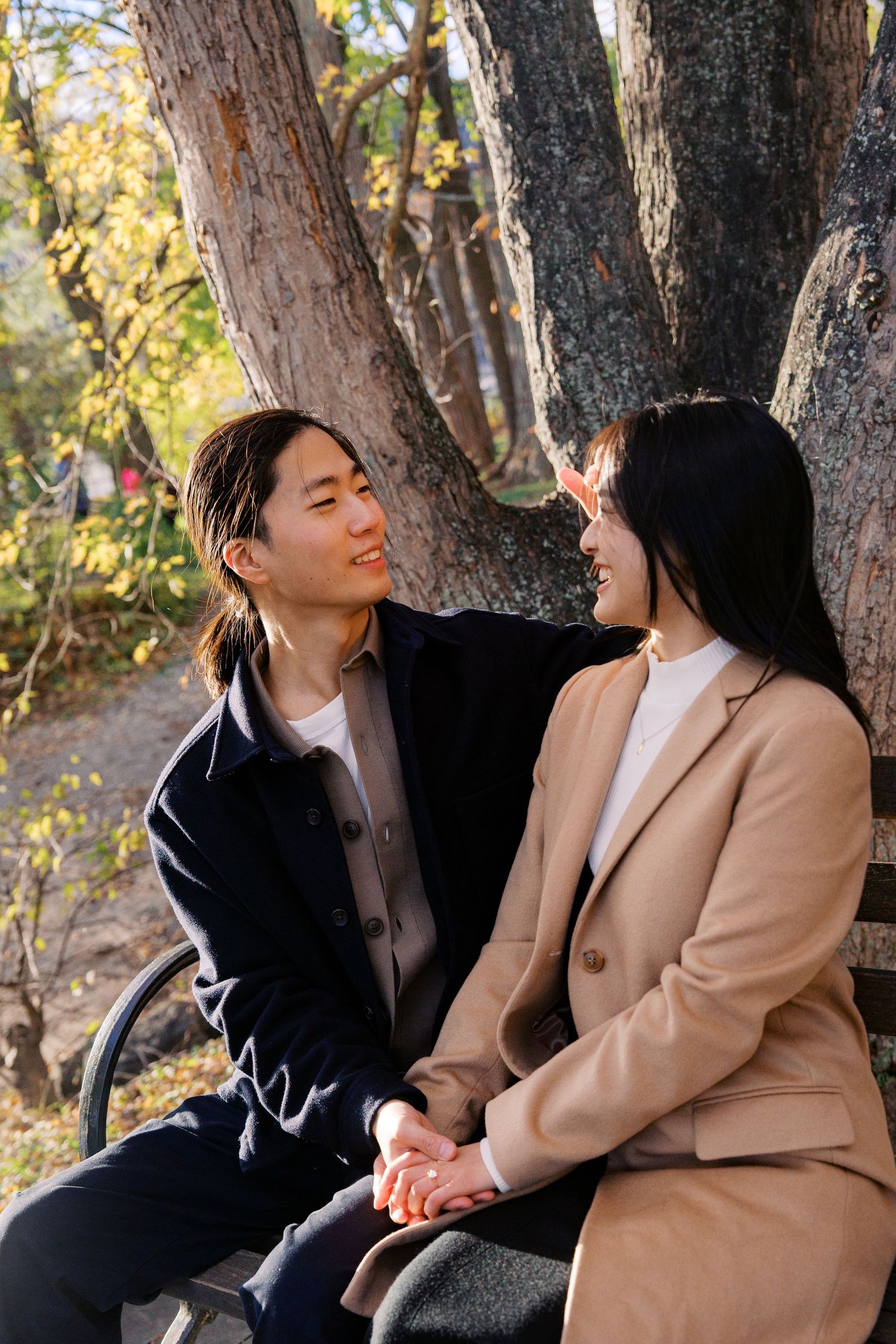 A couple sits together on a bench in a park during autumn, sharing a tender moment among trees.