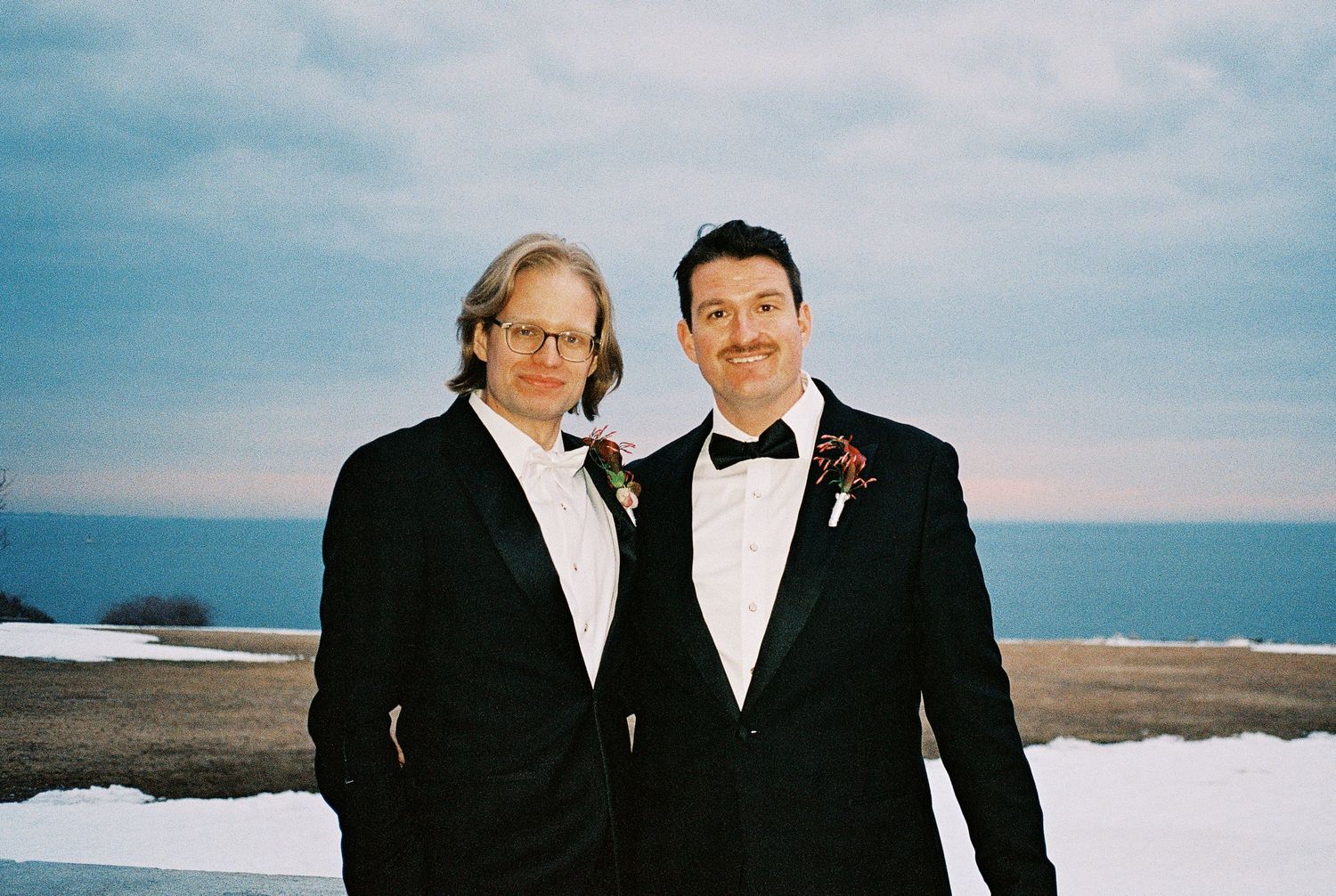 Two people in formal black and white attire stand together against a cloudy sky at the beach.