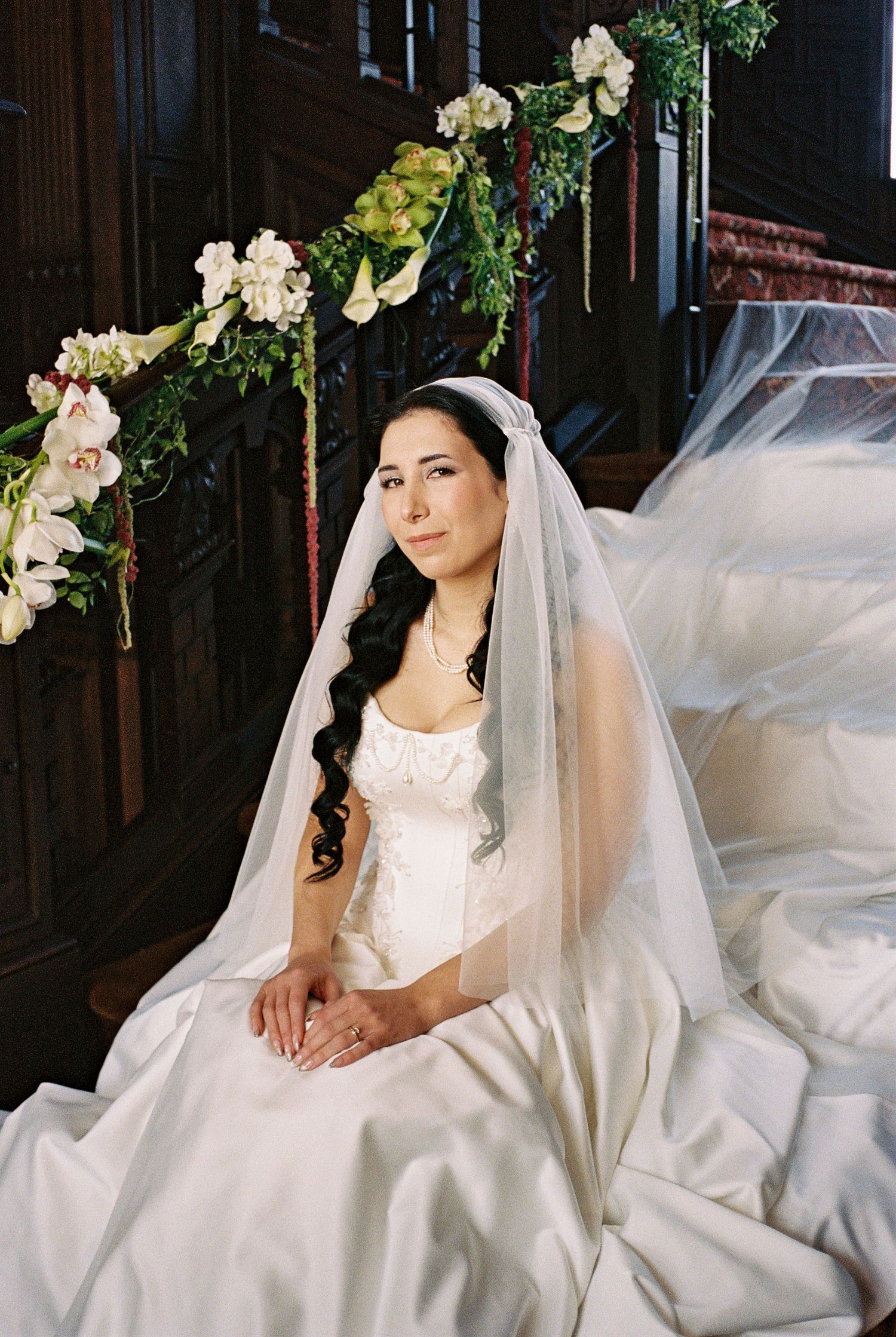 A bride in a flowing white wedding gown and veil poses gracefully against a dark wooden backdrop with floral decorations.