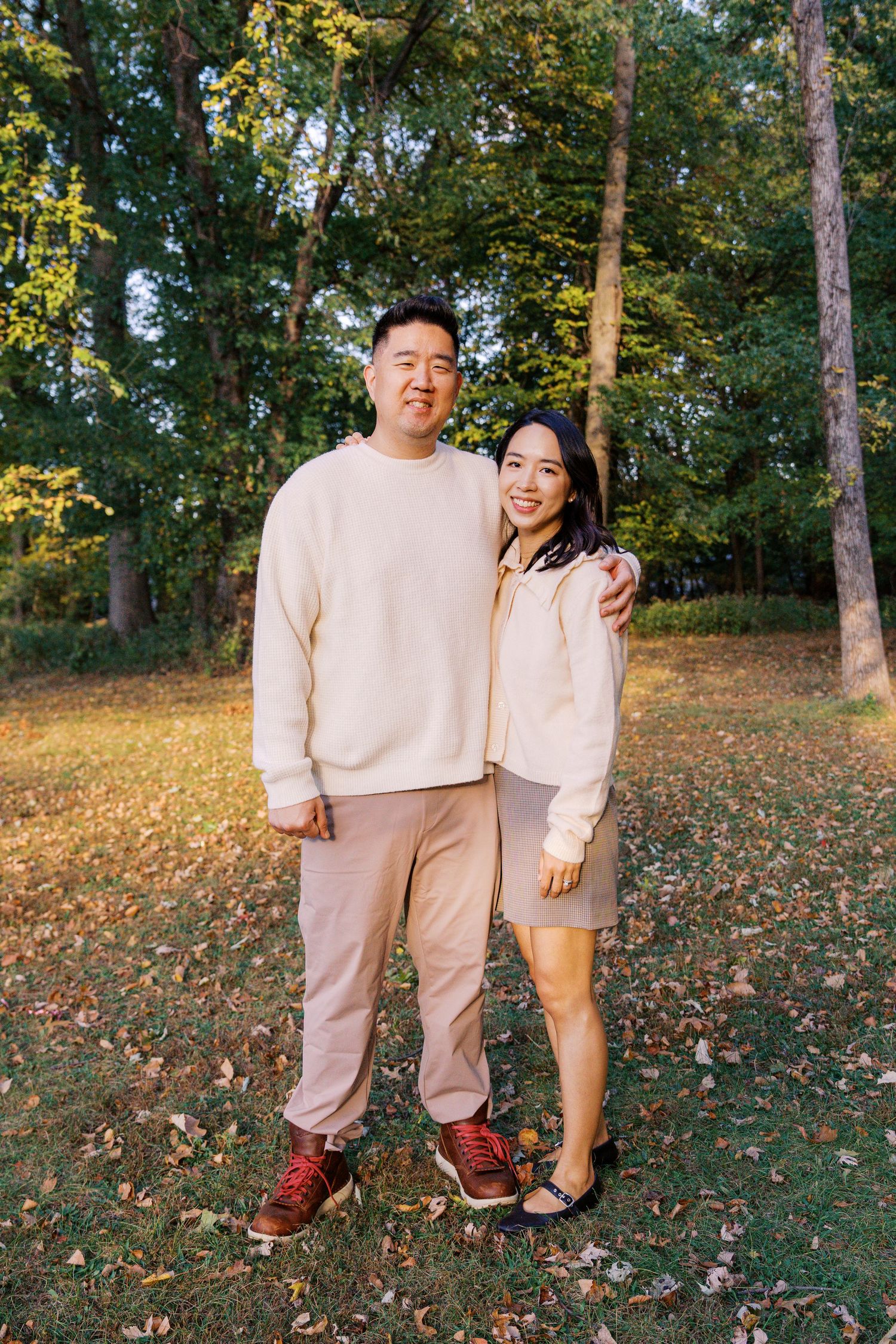 A couple in matching cream outfits poses playfully in a fall forest setting.