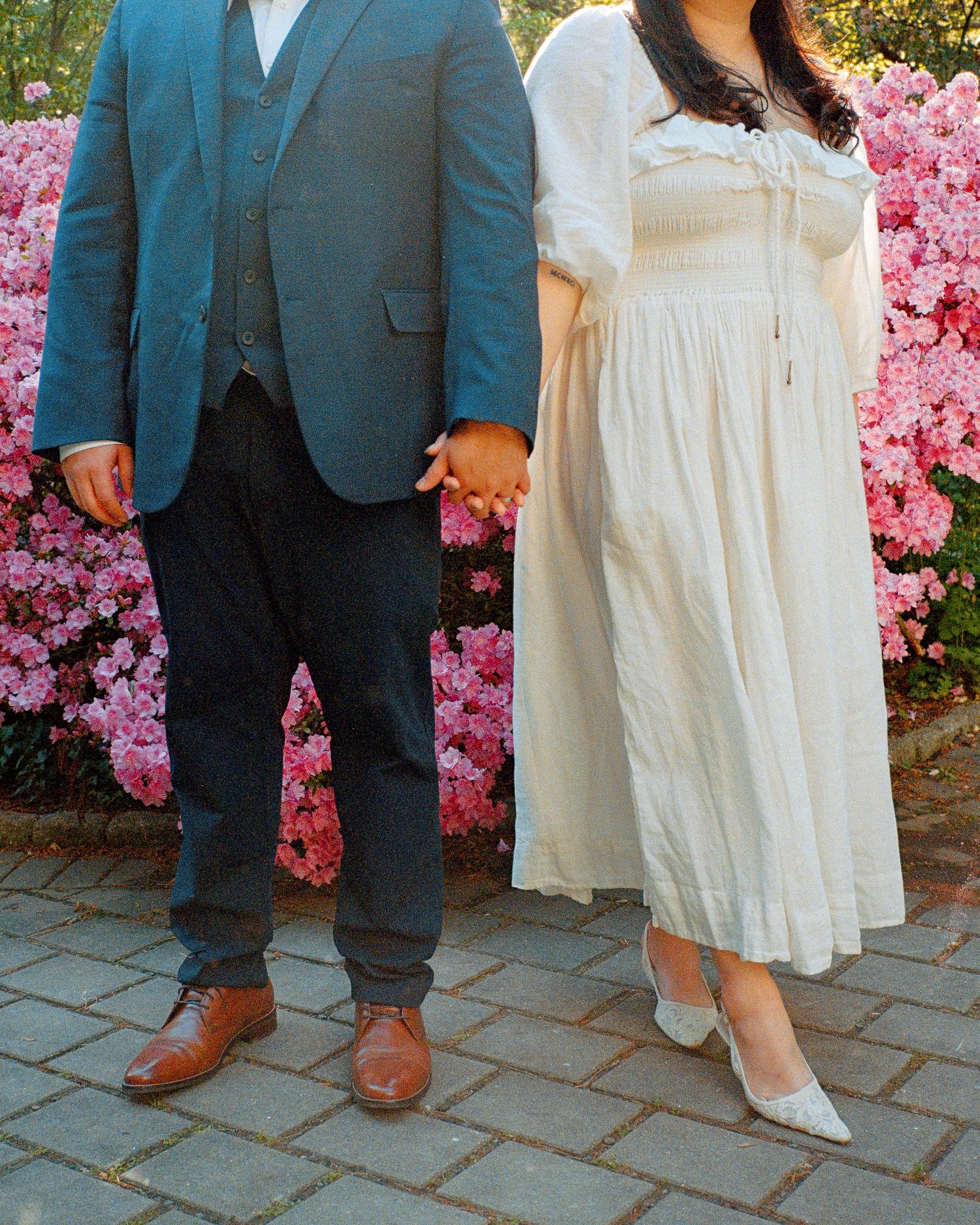 Formal couple poses against blooming pink azalea bushes in garden setting.