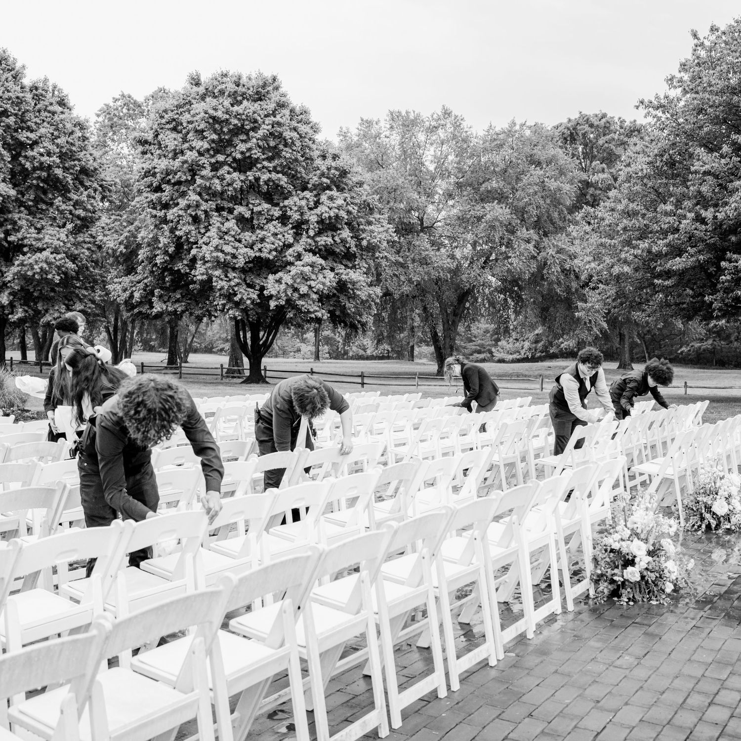 Black and white photo of wedding staff arranging white chairs in rows for an outdoor ceremony surrounded by trees.