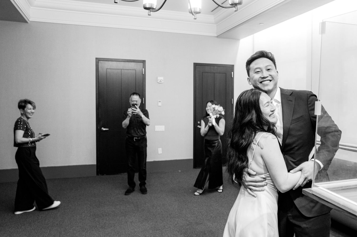 Black and white photo of a couple embracing at a wedding while guests photograph the moment in an elegant venue.