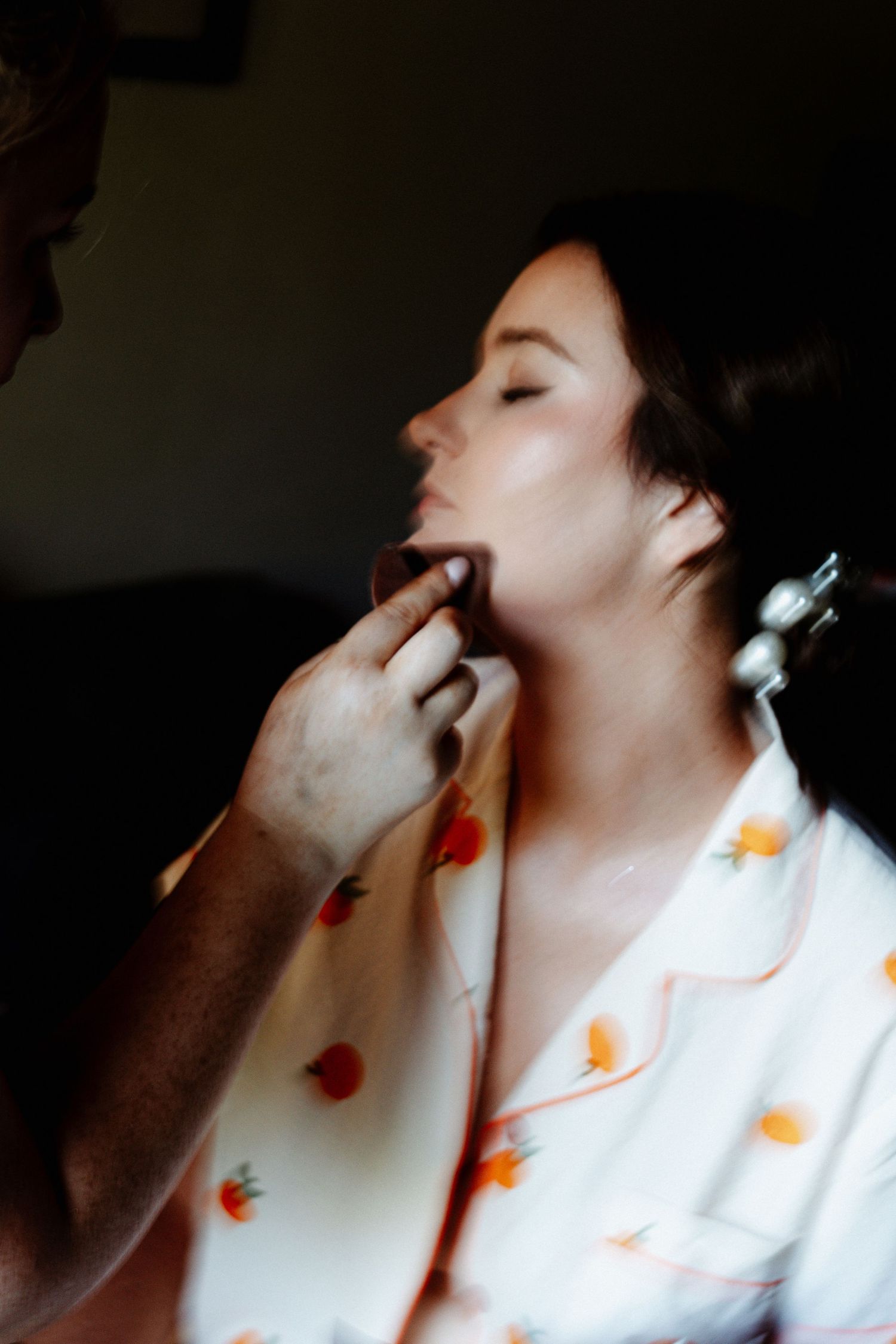 A makeup artist applies foundation to a woman in a floral robe with her eyes closed during bridal preparations.