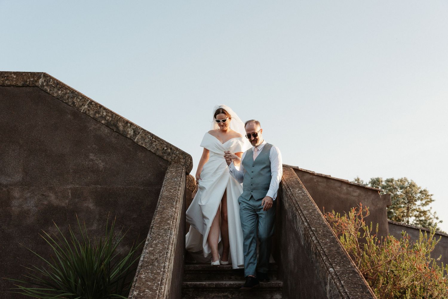 Bride and groom walking down stone steps outdoors, bride in white gown, groom in grey suit, blue sky background.