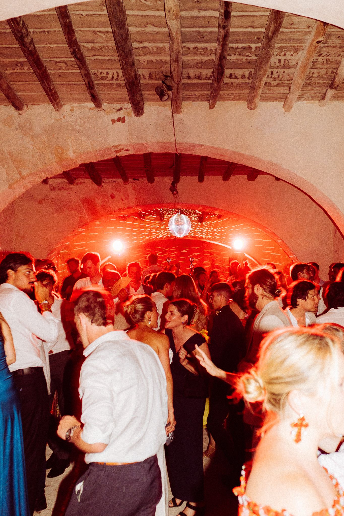Crowded wedding dance floor with red lighting in rustic arched venue.