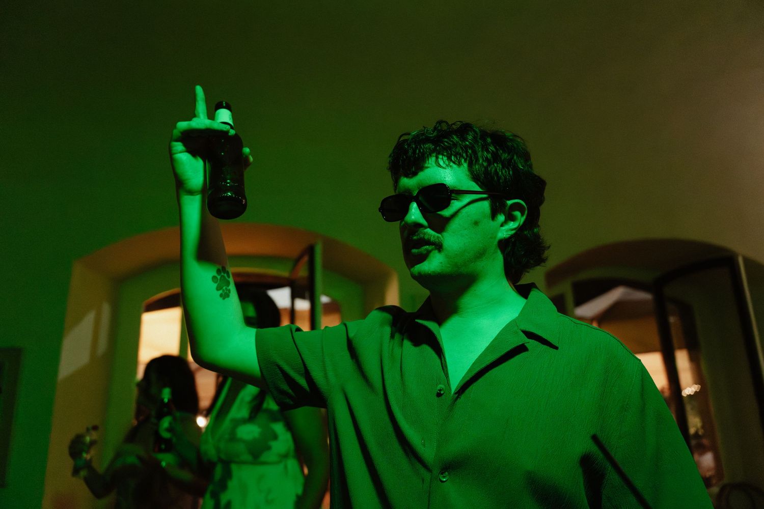 Man in green shirt with sunglasses holding drink under green lighting at a party.