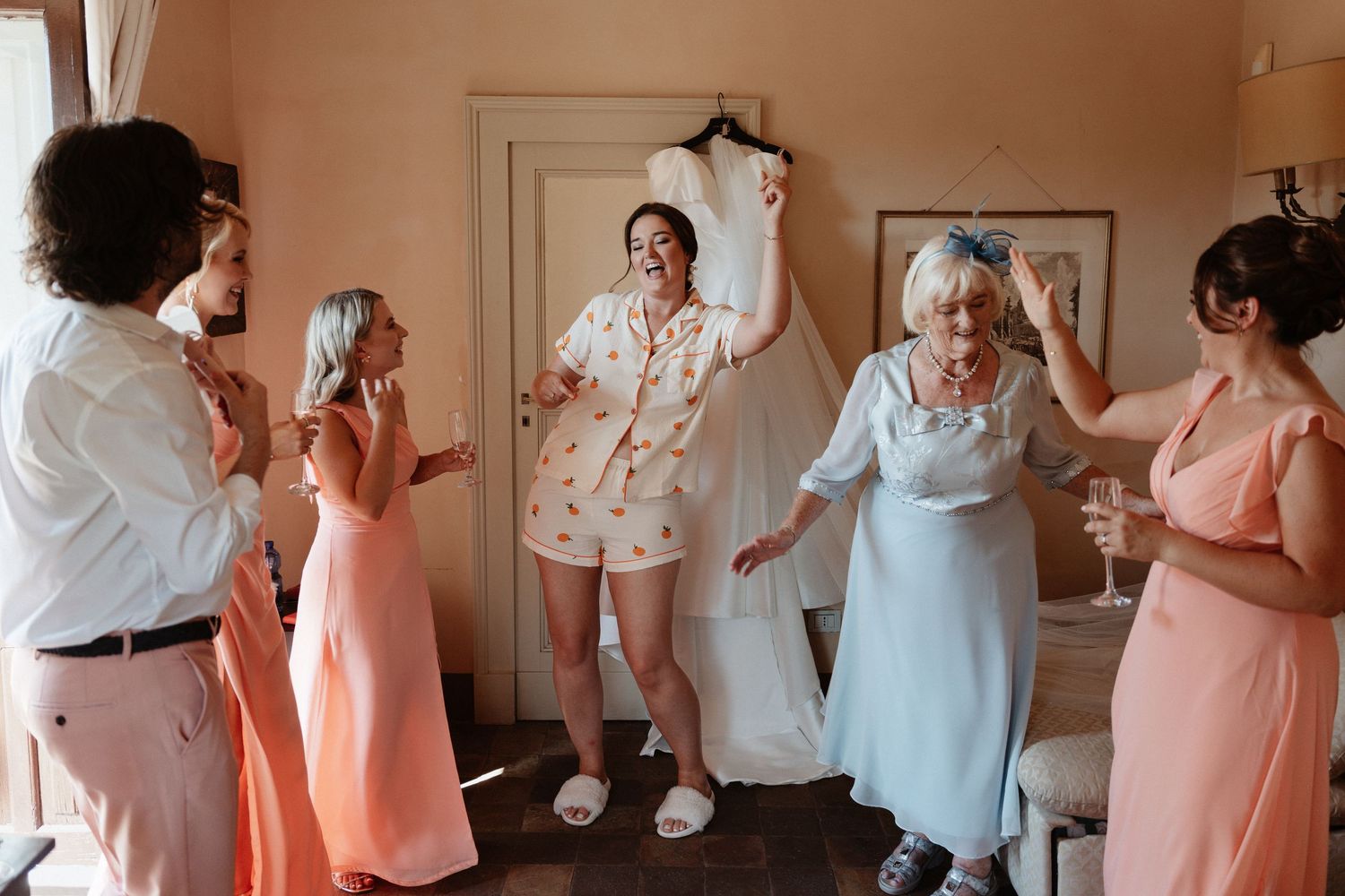 Bride in pajamas dances joyfully with bridesmaids in pink dresses and elderly guest, wedding gown hanging in background.