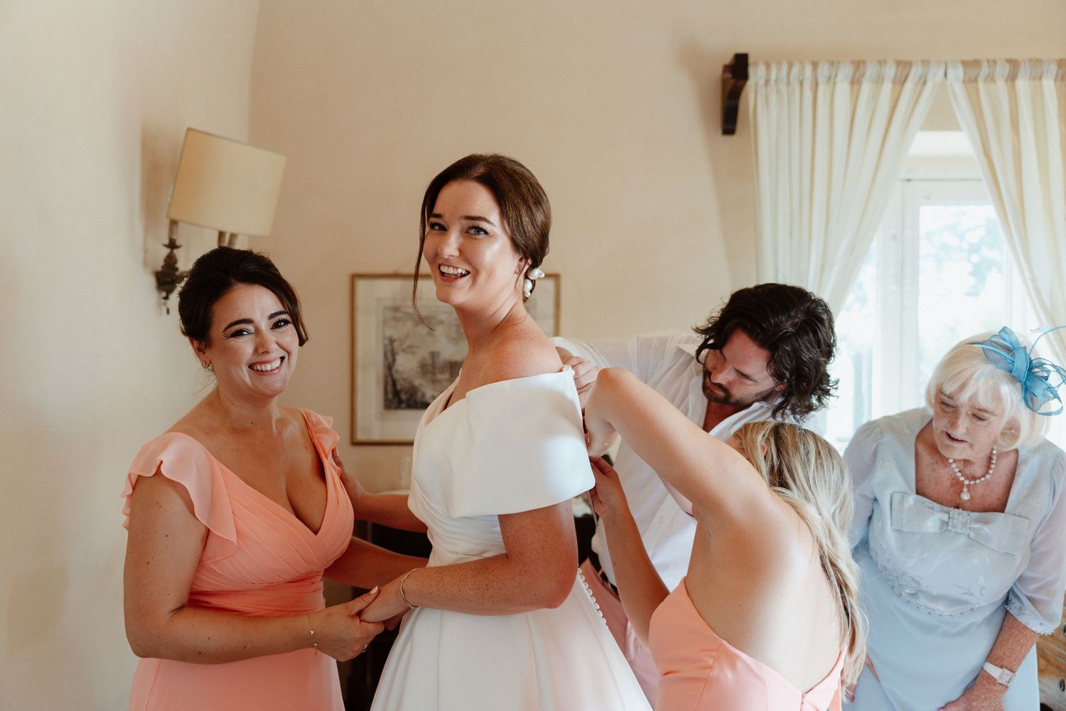 Bride in white off-shoulder gown being helped by bridesmaids in pink dresses during wedding preparation indoors.