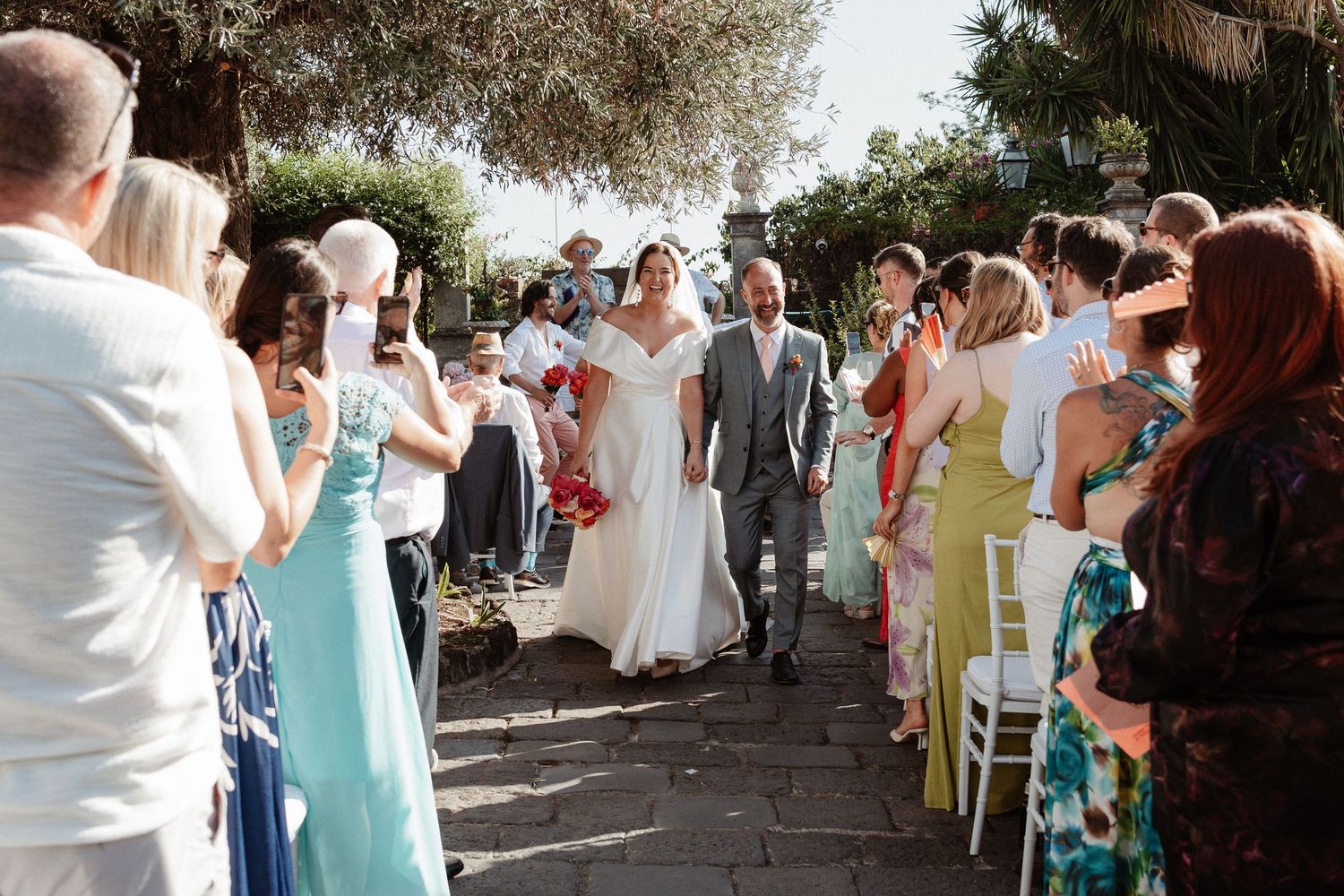 Bride and groom walk down the aisle together surrounded by smiling wedding guests at an outdoor ceremony.
