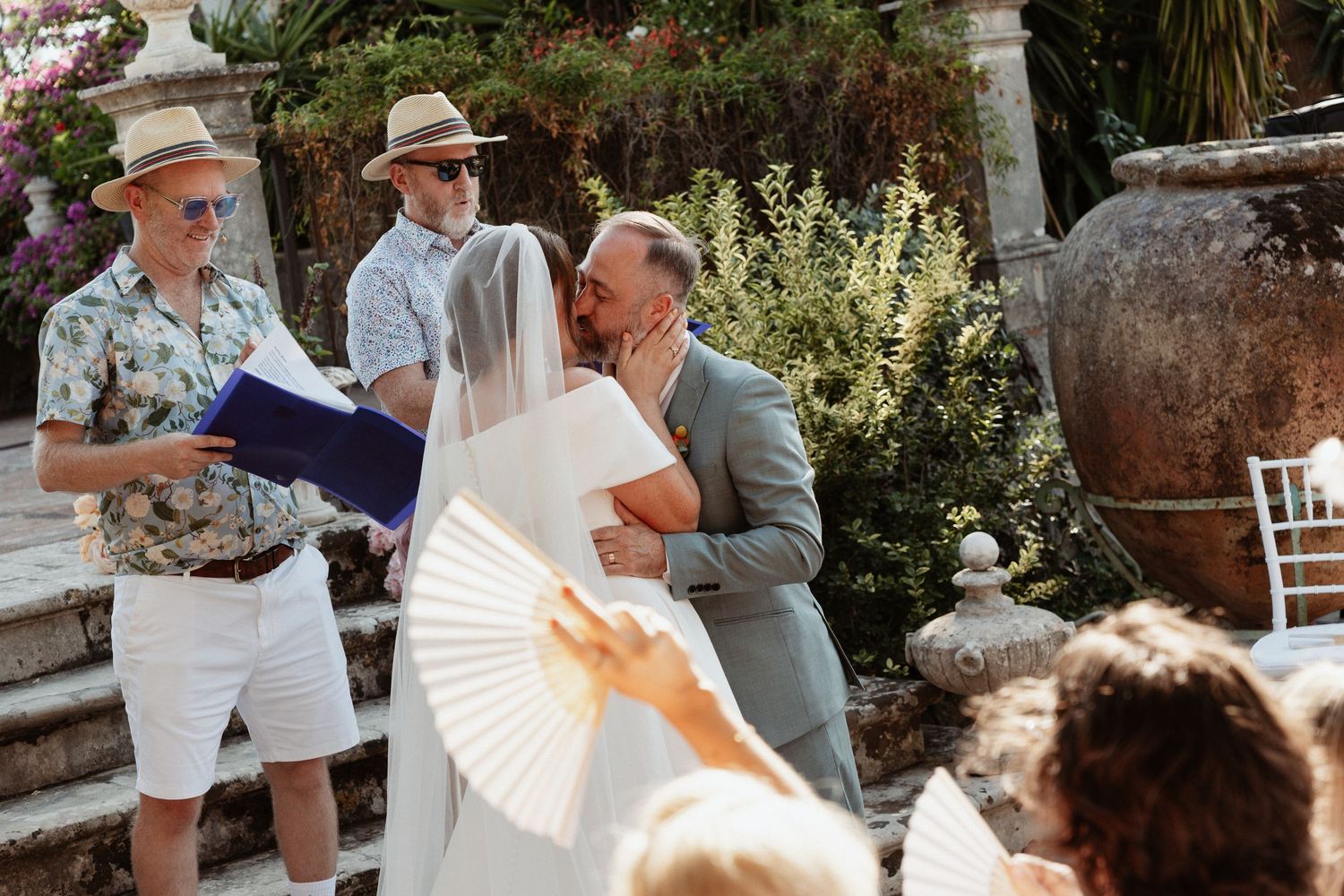 Bride and groom share a kiss at an outdoor wedding ceremony while guests watch and an officiant holds a blue book.