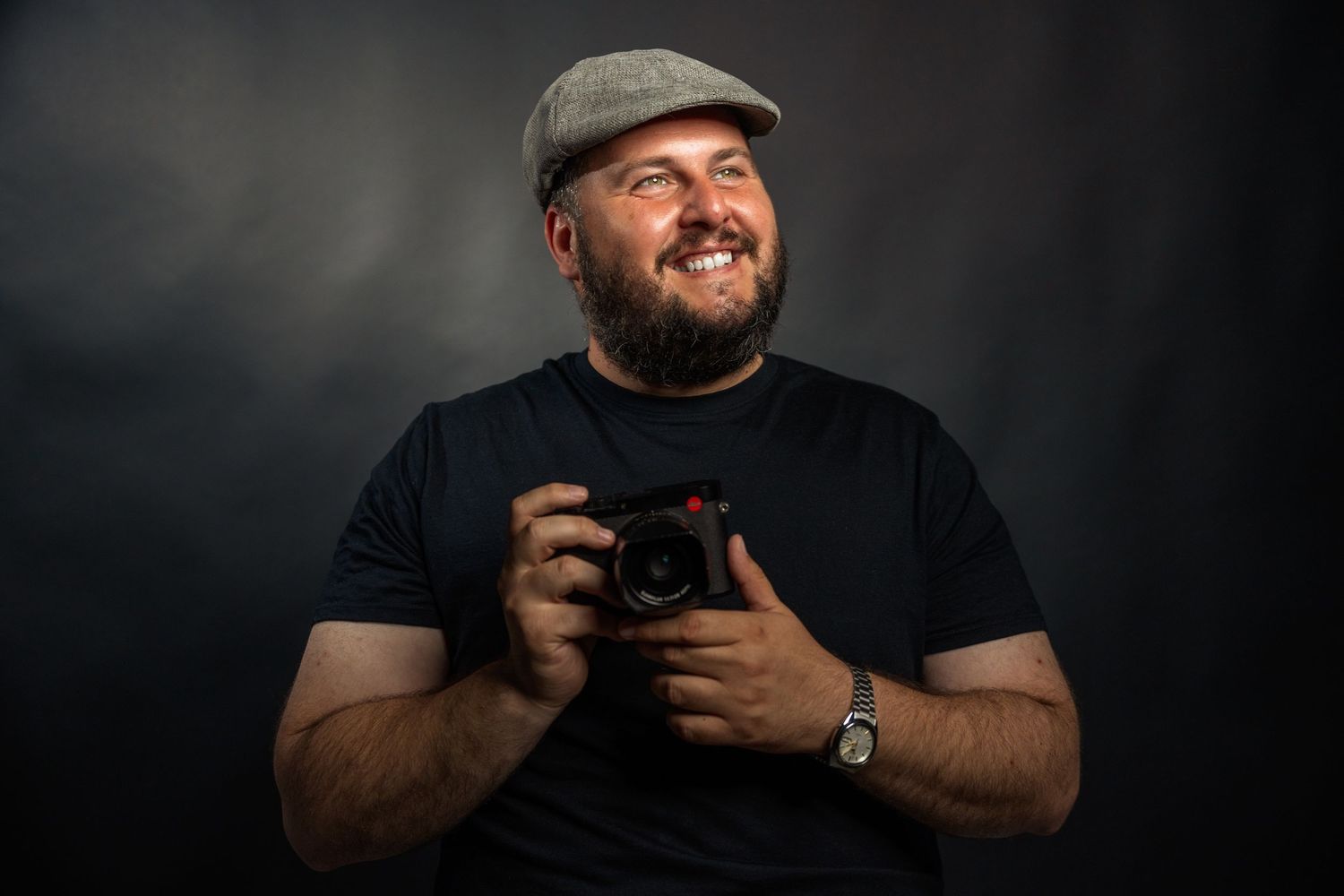 Cheerful photographer in cap holding camera against dark studio backdrop.