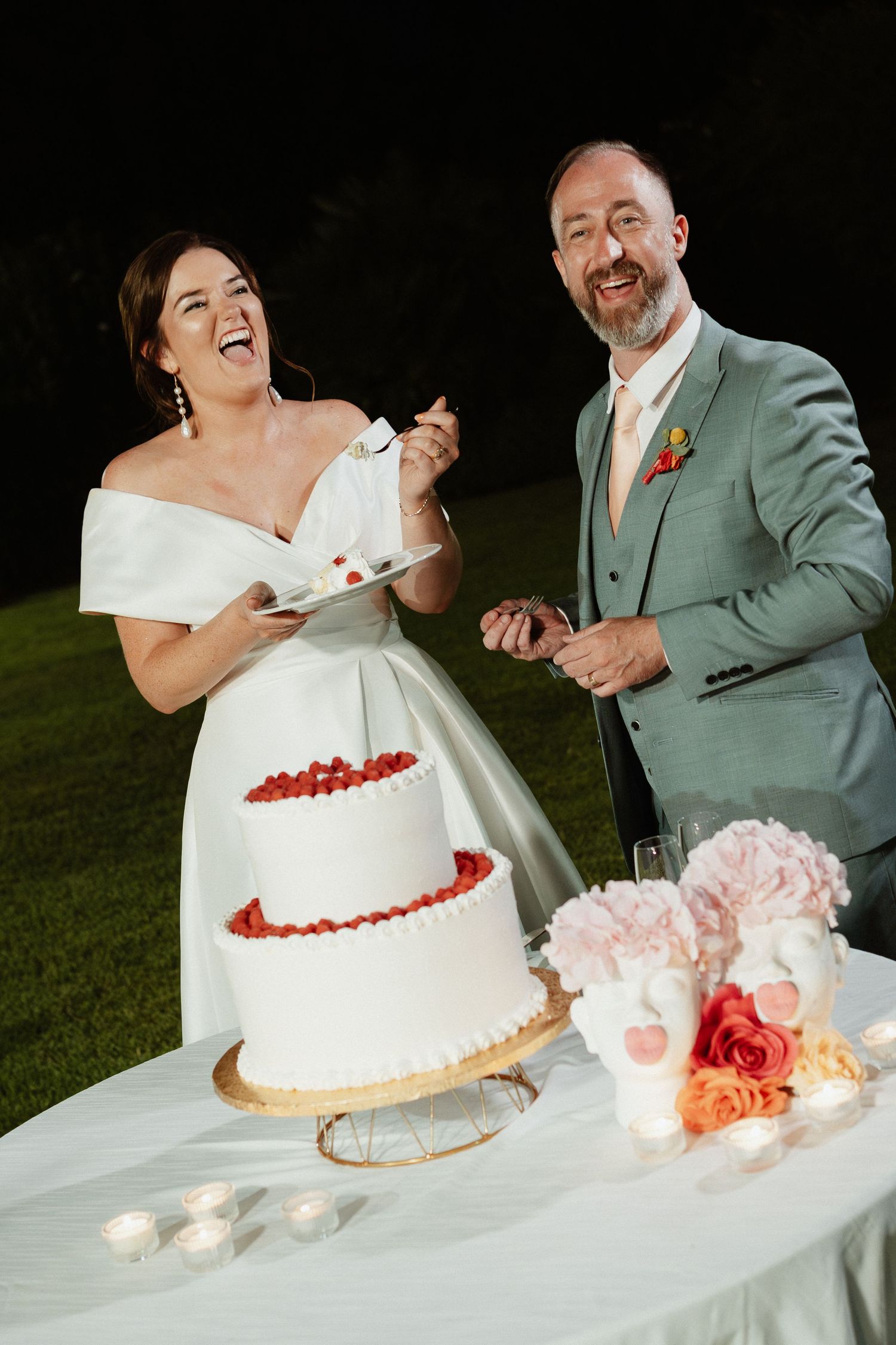 Bride and groom laughing while cutting a two-tier white wedding cake decorated with fresh strawberries outdoors at night.