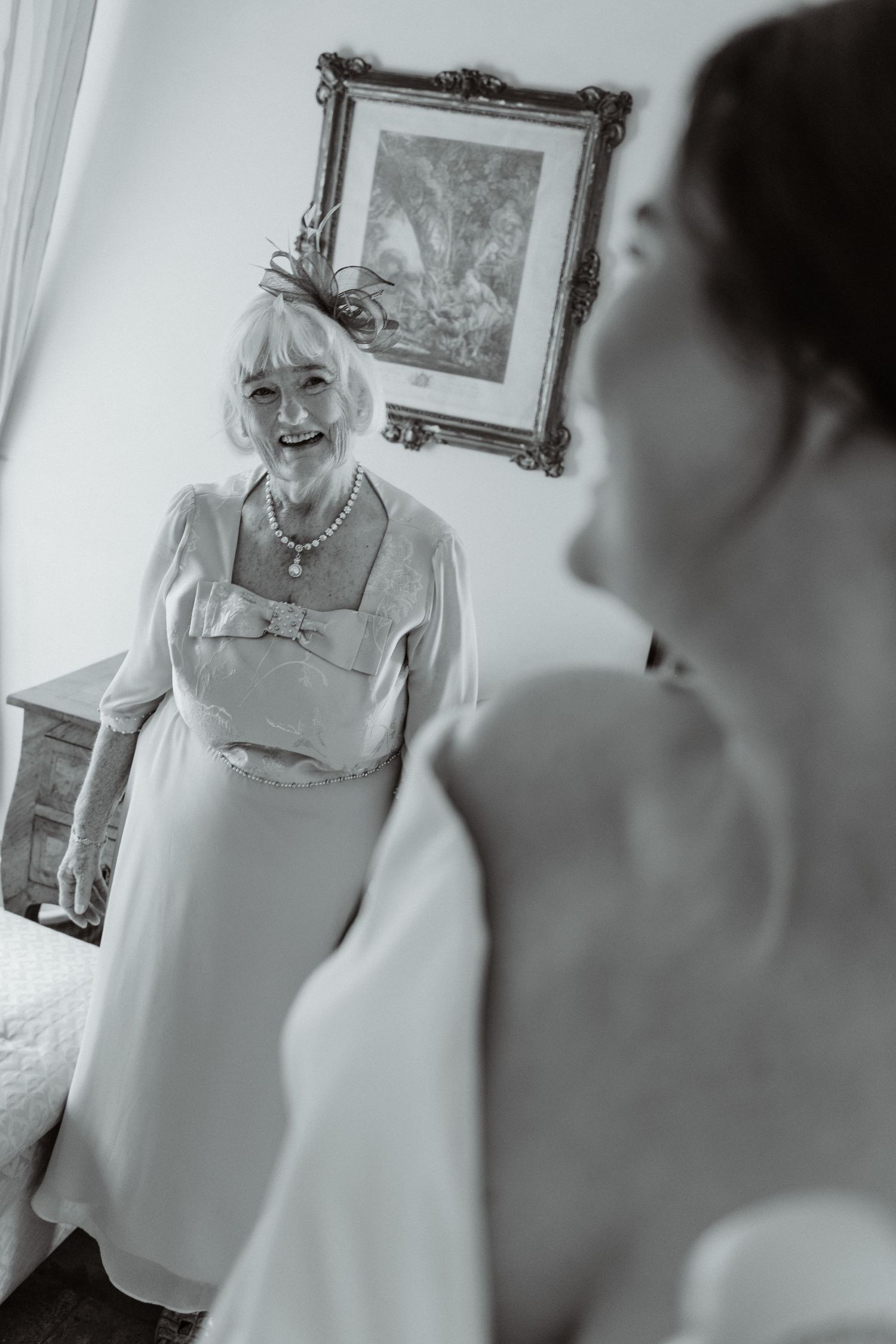 Black and white photo of smiling elderly woman in elegant dress watching bride in wedding gown during bridal preparation.