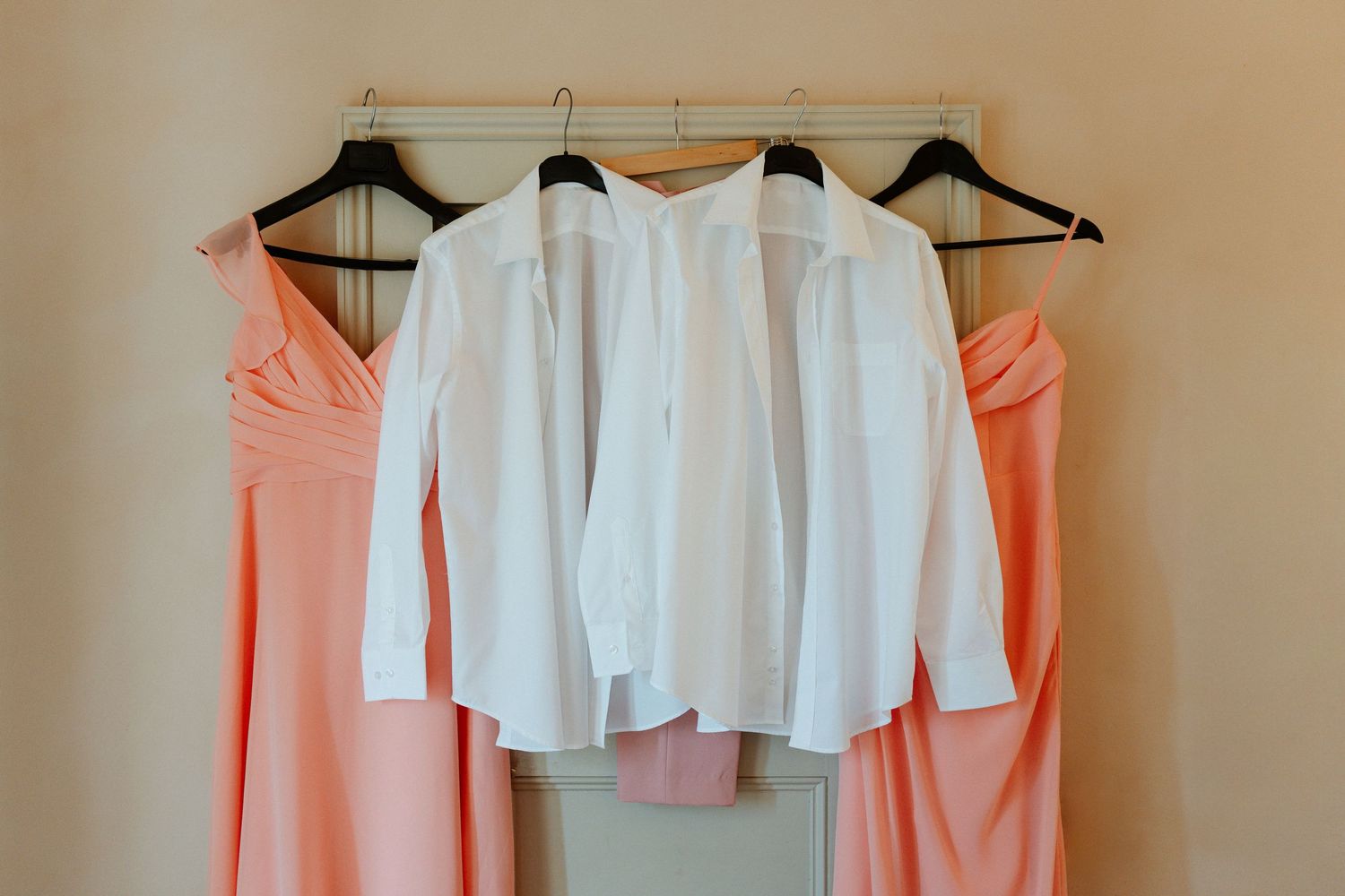 Two peach bridesmaid dresses and two white dress shirts hanging on black hangers against a neutral wall.