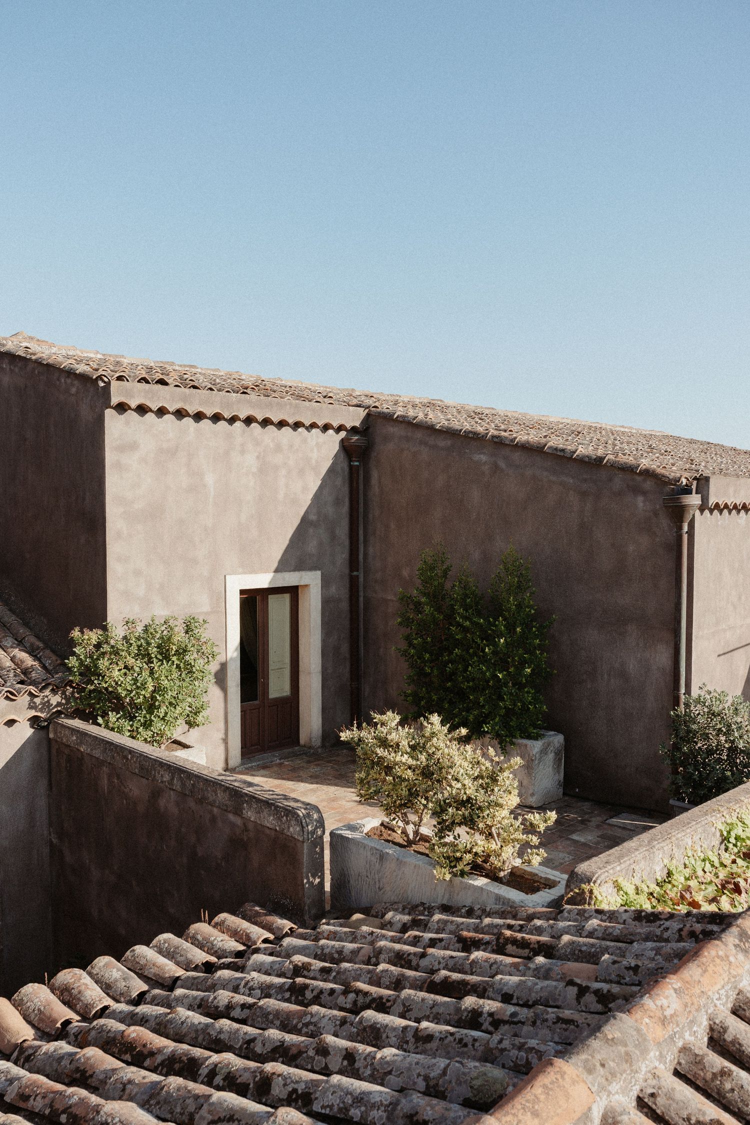 Rustic Mediterranean rooftop with terracotta tiles, weathered stone walls, green plants, and a wooden door under clear blue sky.