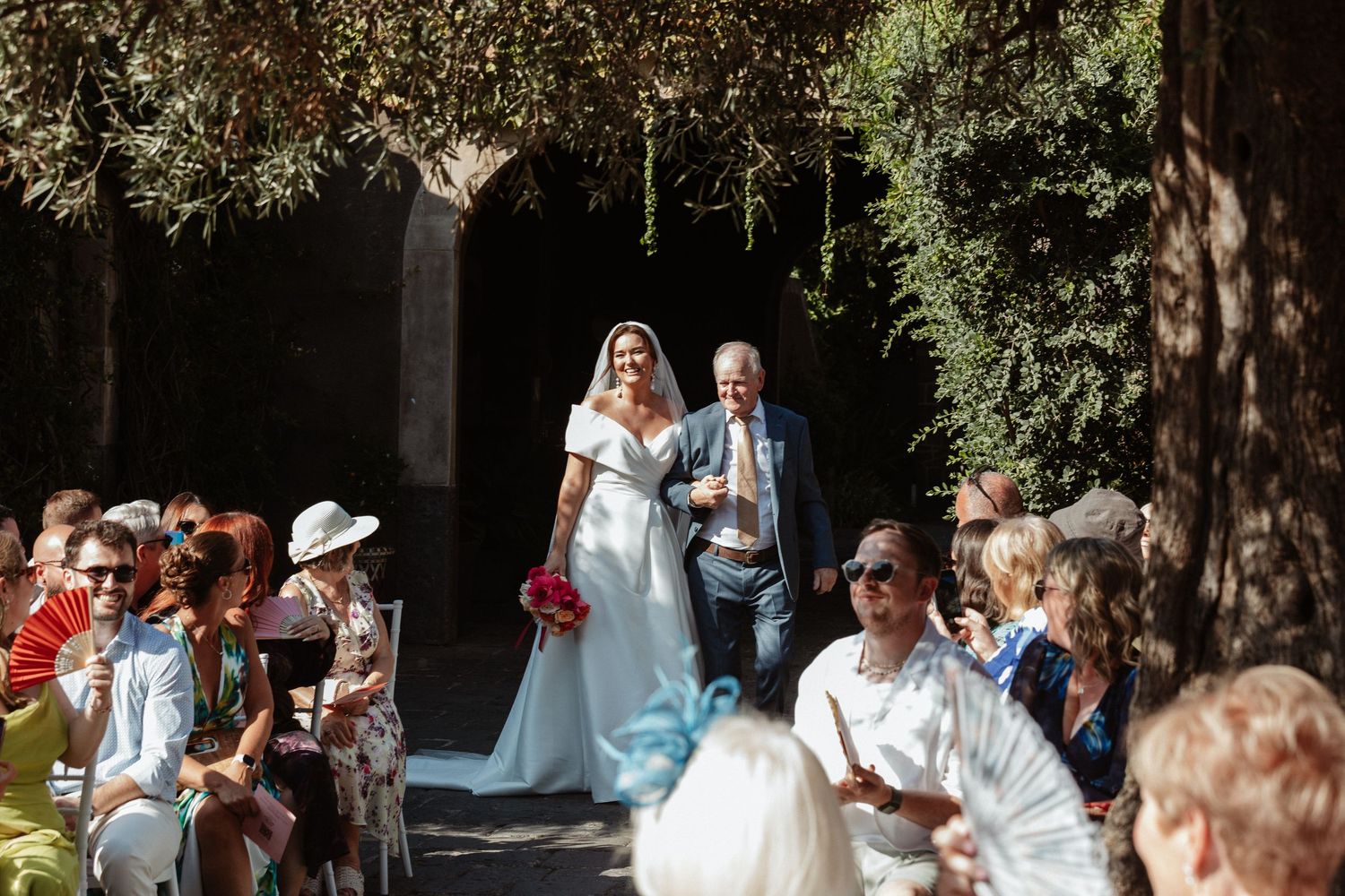 Bride in white gown walks down outdoor aisle with escort at sunny garden wedding ceremony.