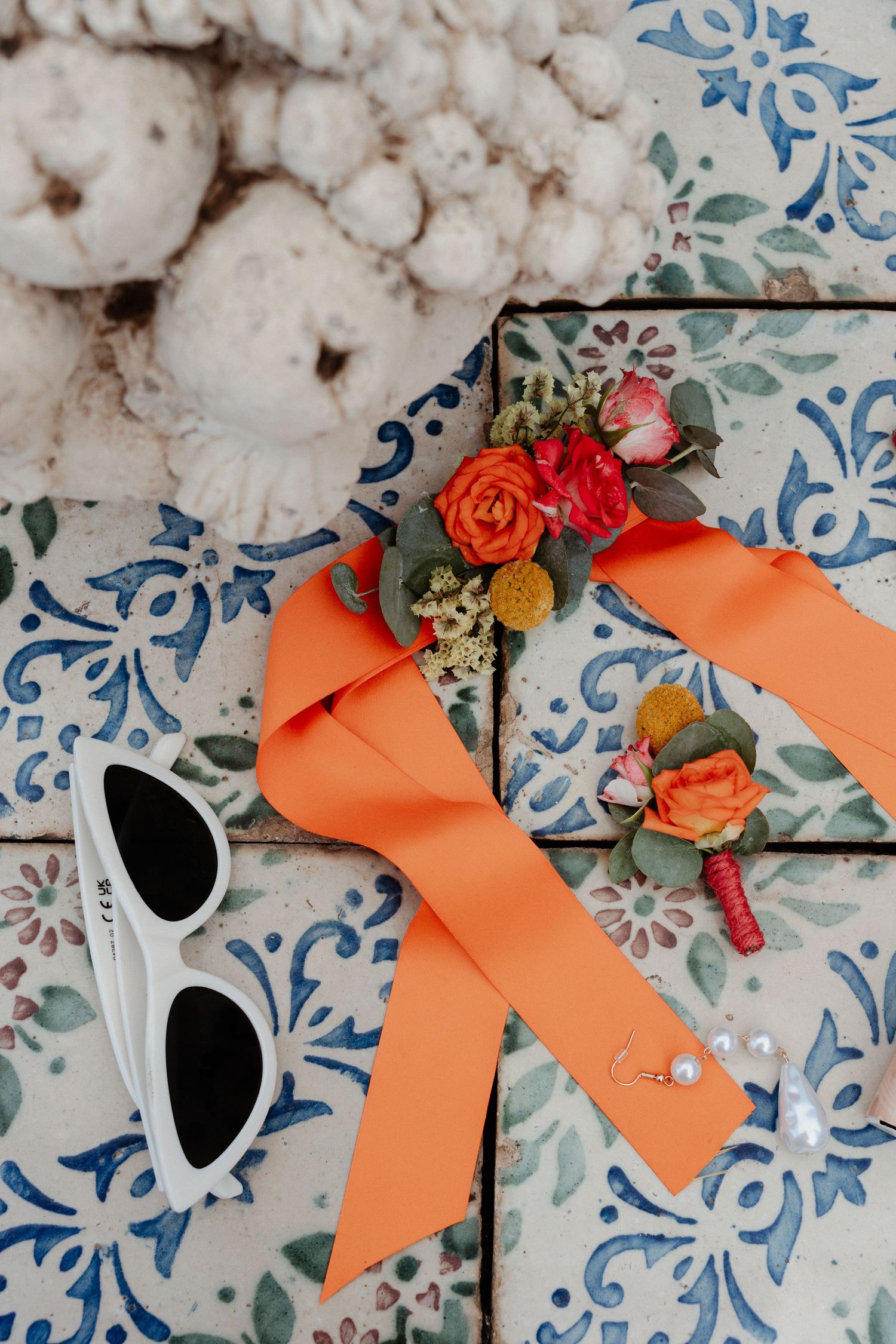 Orange ribbon boutonniere with colorful flowers beside white sunglasses on decorative blue tile background.