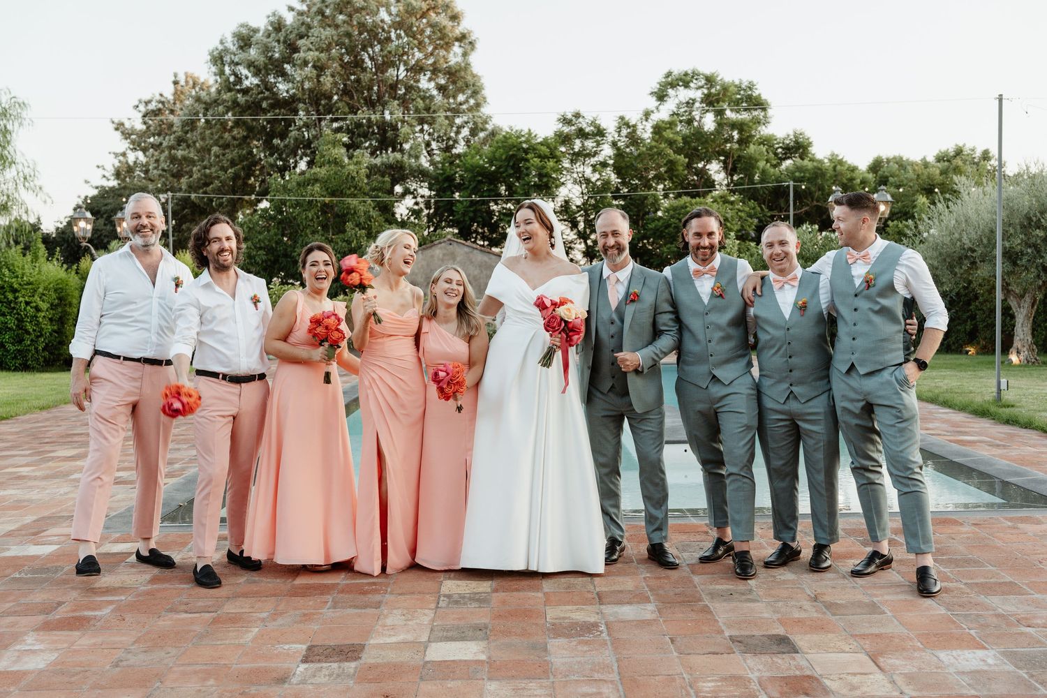 Wedding party posing outdoors, bride in white gown with bridesmaids in coral dresses and groomsmen in gray suits.