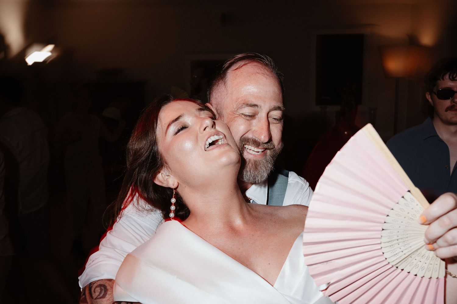 Bride and older man laughing joyfully at wedding reception, holding a pink decorative hand fan.
