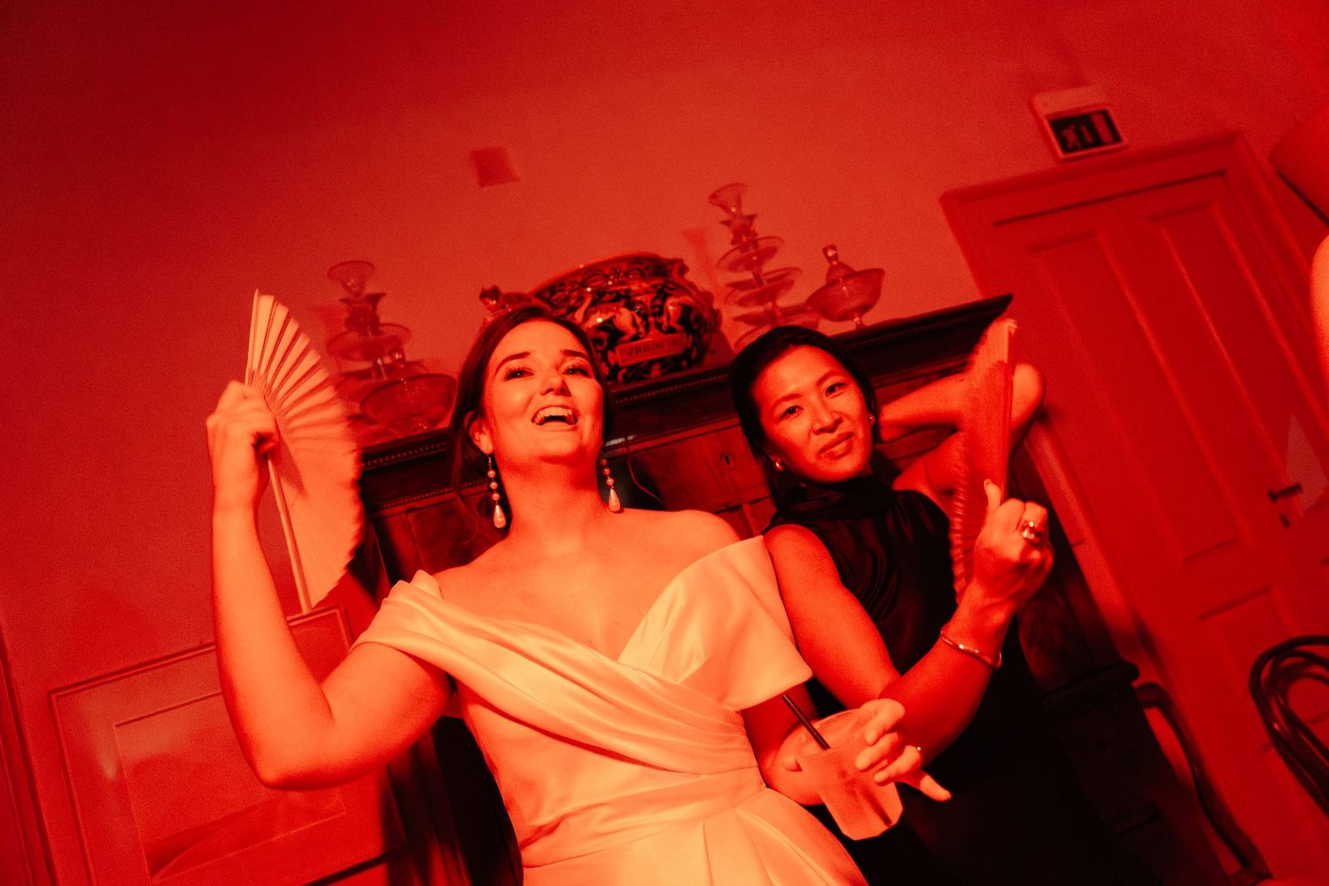 Two women dancing joyfully under red lighting, one in a white dress holding a decorative fan at an elegant party.