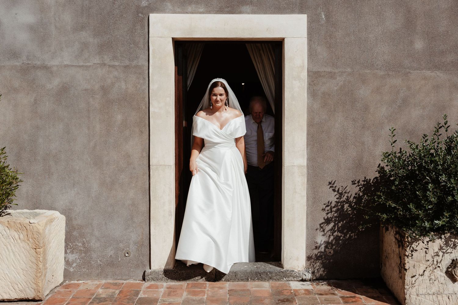 Bride in white off-shoulder gown and veil standing in stone doorway at elegant outdoor wedding venue.