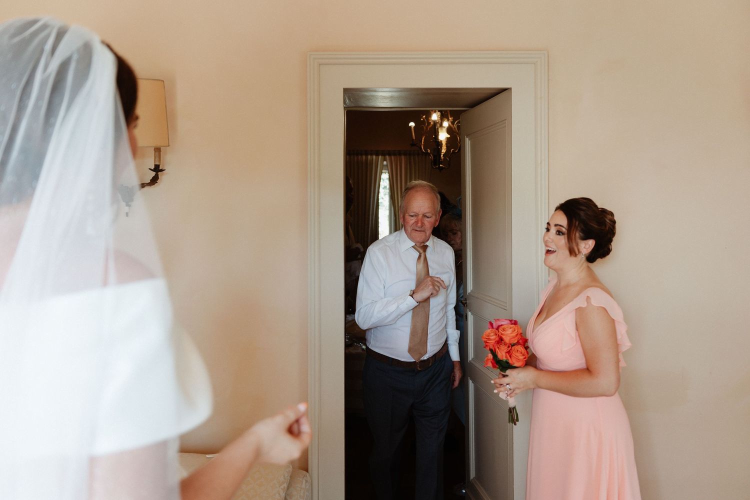 Father of the bride smiles with emotion as he sees his daughter in her wedding dress, bridesmaid holding bouquet nearby.