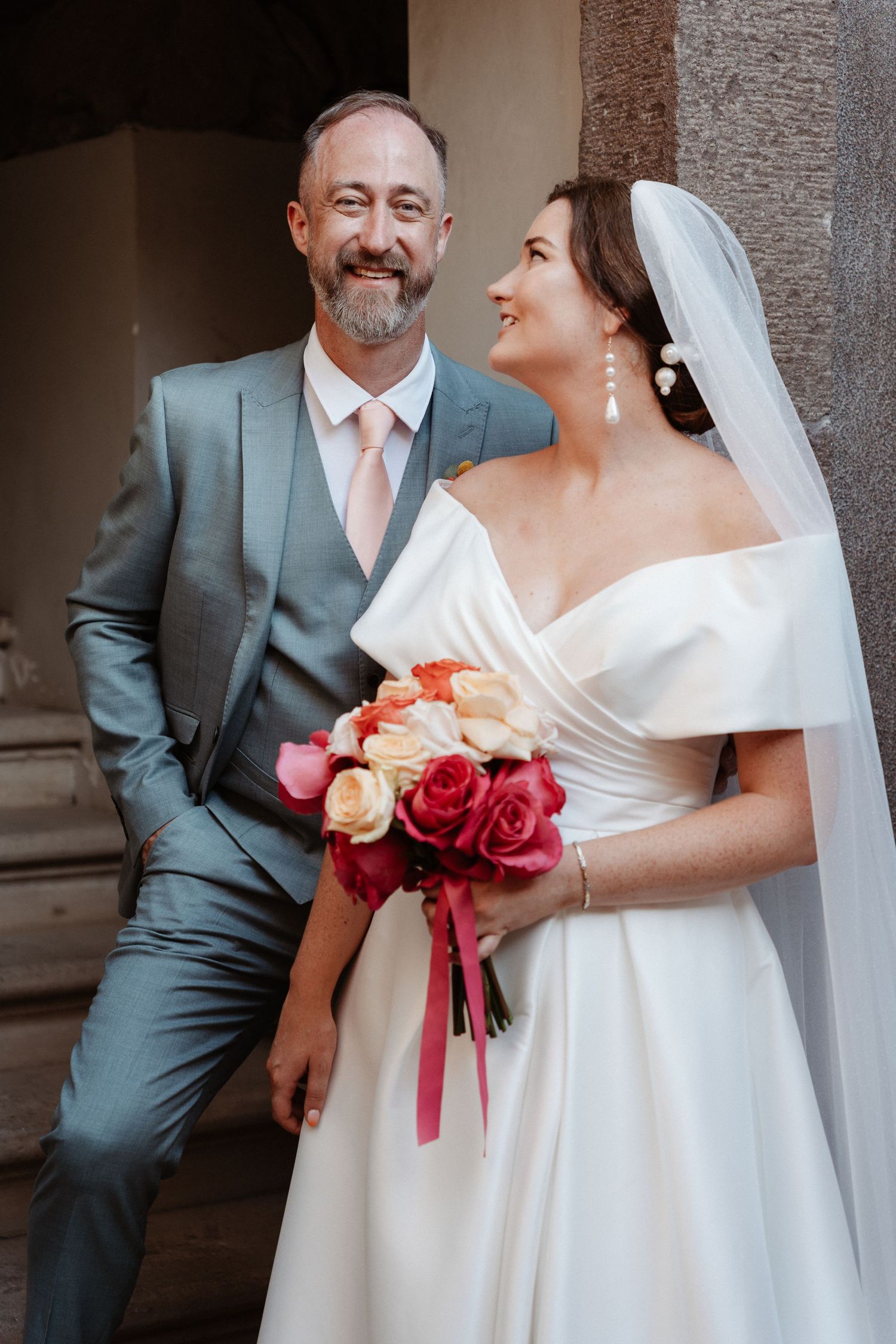 Bride in white off-shoulder gown holding colorful rose bouquet smiles at bearded groom in gray suit with pink tie.